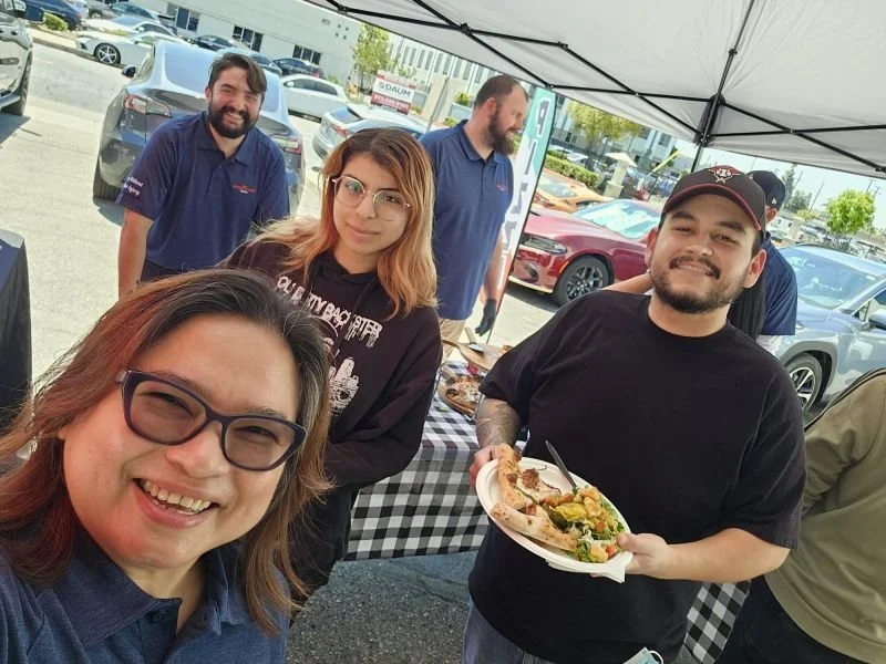 Group of five people at an outdoor gathering, with two men in blue shirts in the background and a woman taking a selfie with a man holding a plate of grilled food in the foreground.