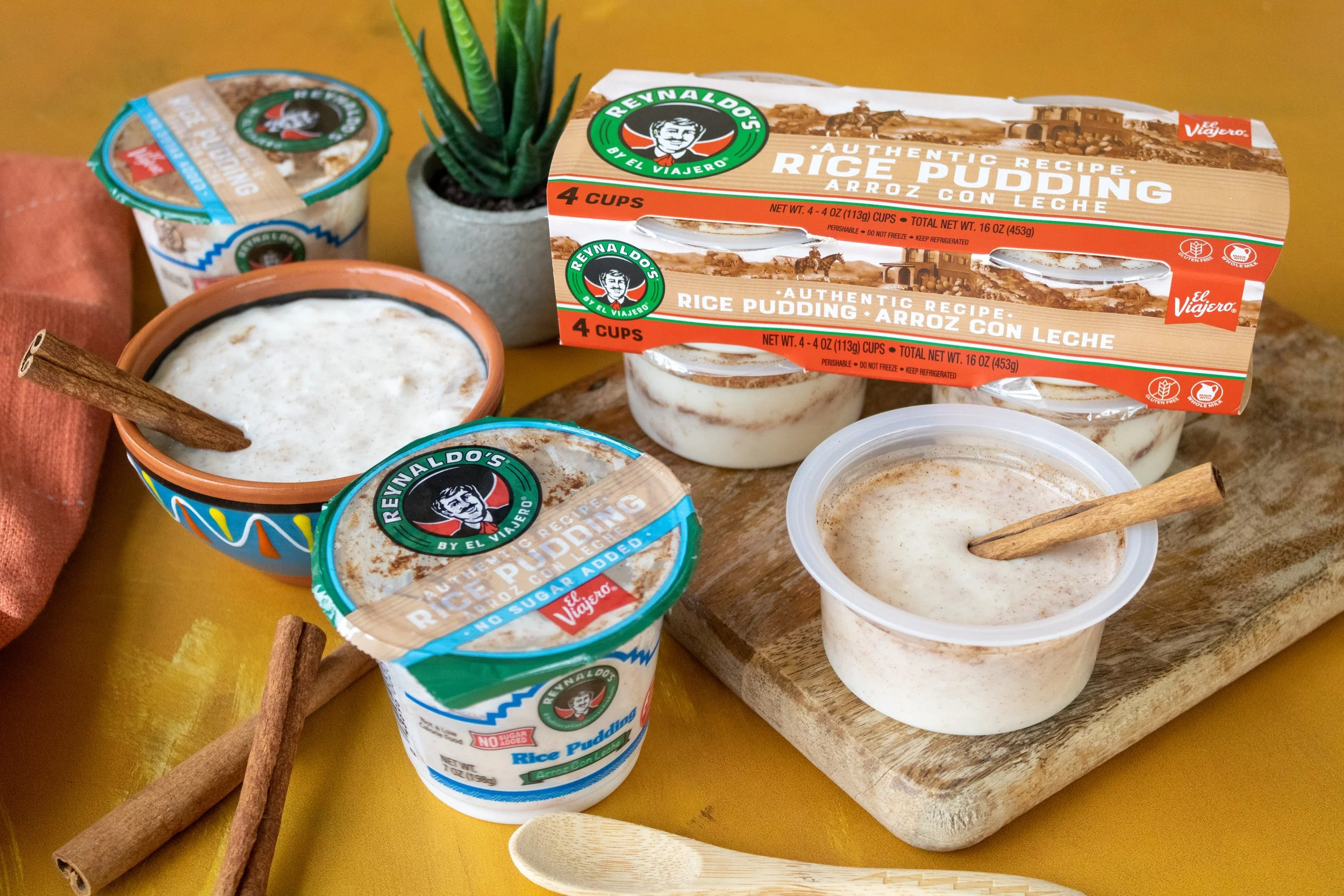 Various containers of rice pudding from Reynaldo's, some with cinnamon sticks, a small potted succulent plant, a wooden spoon, and a cloth, all on a yellow surface.