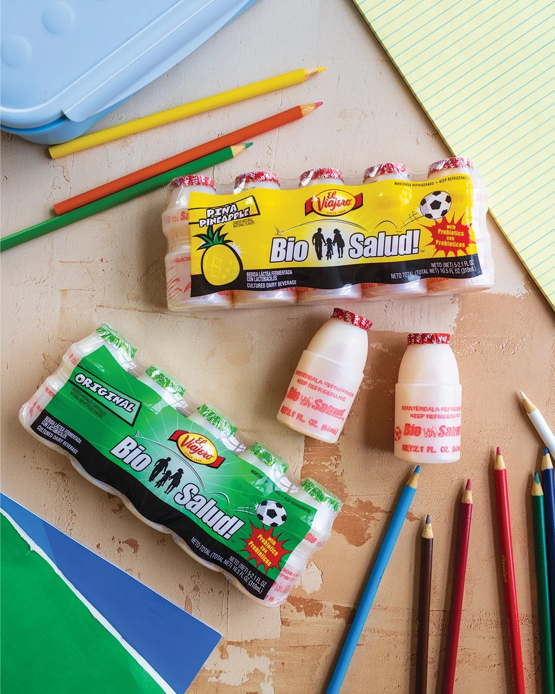 Two packs of probiotic yogurt bottles, colorful pencils, a yellow notebook, and a blue box on a wooden surface.