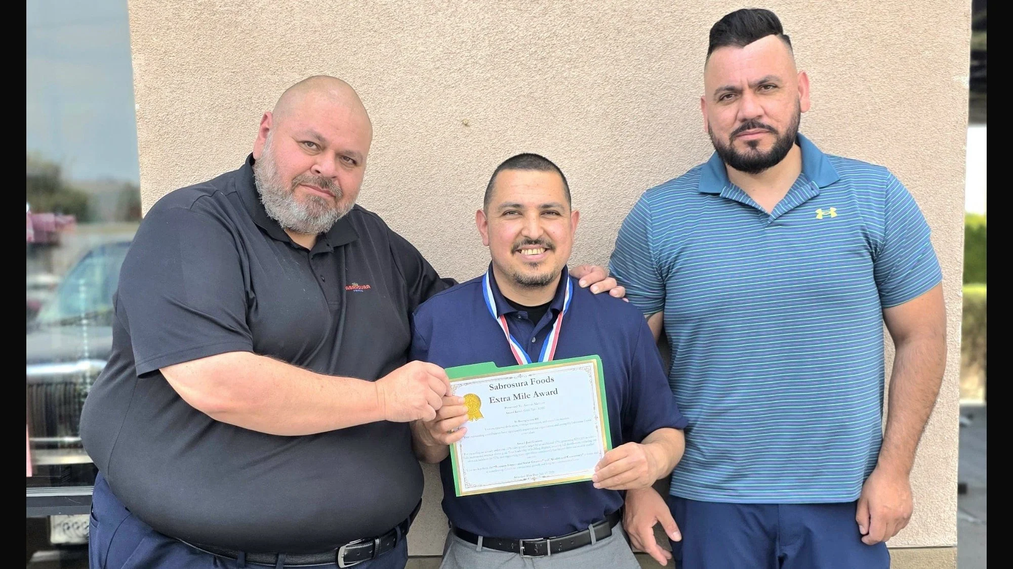 Three men standing together outdoors in front of a beige wall. The man in the middle is holding a certificate that reads "Sabrosura Foods Extra Mile Award" and is wearing a medal around his neck. The other two men are standing on either side of him,