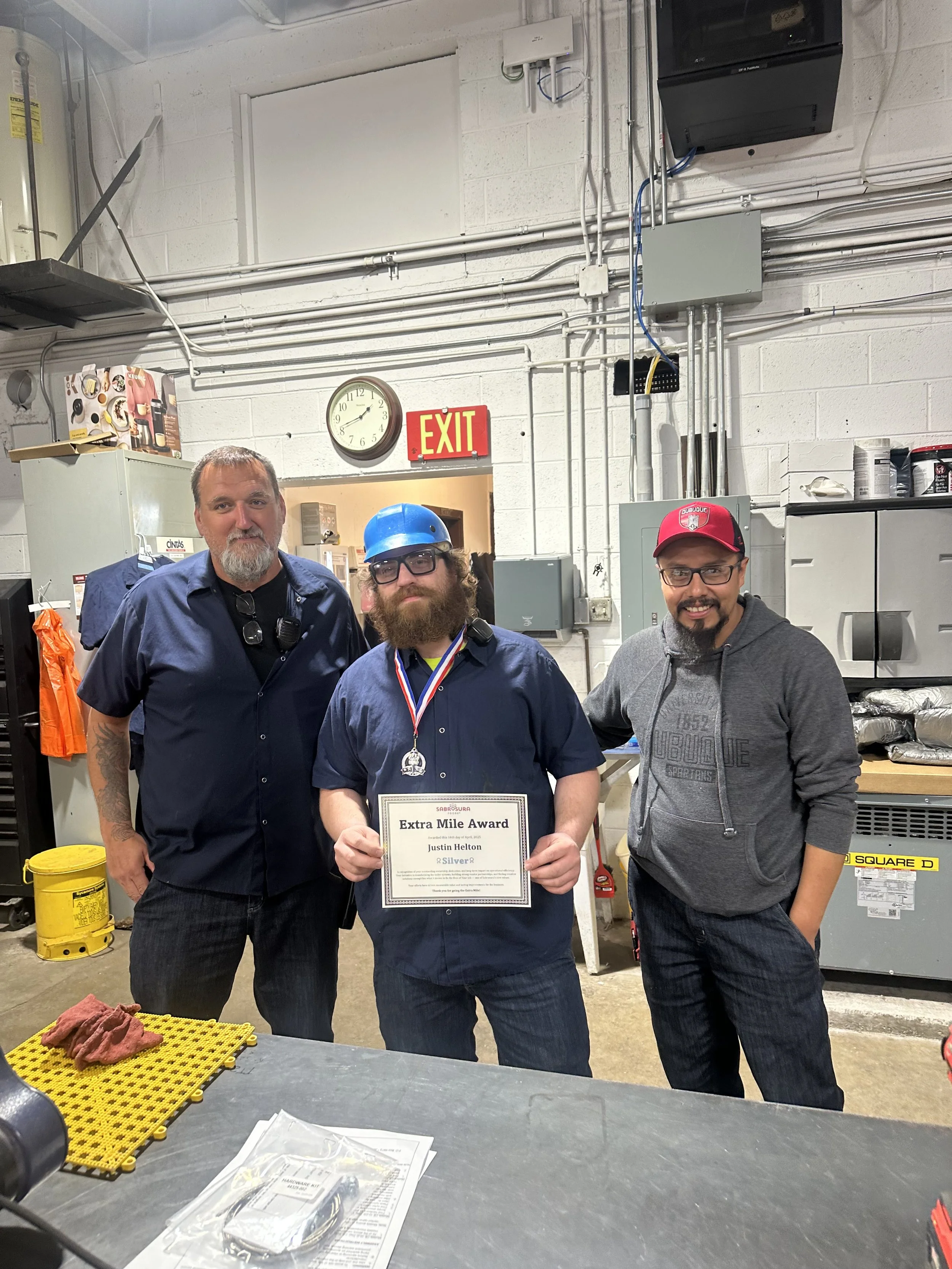 A man wearing glasses and a blue helmet holding an award certificate, standing between two men, in a workshop or industrial setting with tools, cabinets, and equipment around.