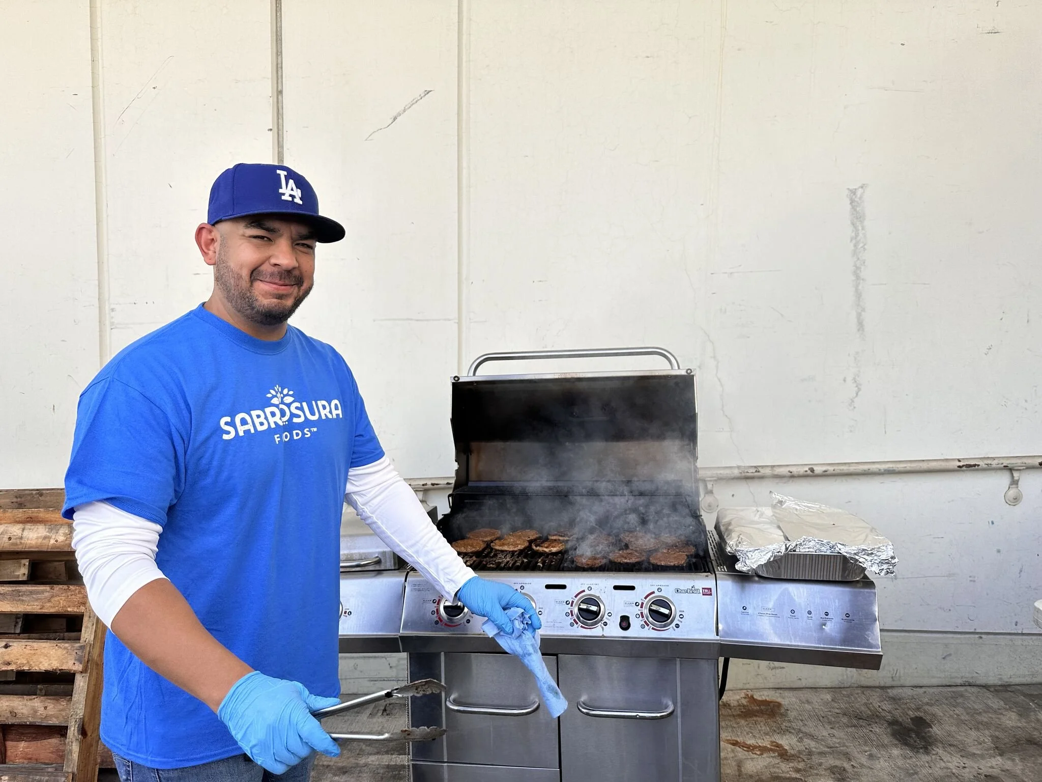 Man grilling hamburgers outside on a stainless steel barbecue grill, wearing a blue Los Angeles Dodgers cap, a blue Sabrosura Foods T-shirt, and blue gloves, smiling at the camera.