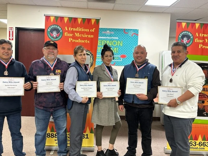 Six people standing side by side holding certificates and medals, in front of branded banners promoting Mexican products at an award ceremony.
