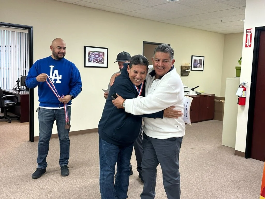 Four men in an office space celebrating. Two men in the center are hugging and smiling, one wearing a white jacket and the other a dark jacket. A man on the left is holding medals, and a man behind them is partially hidden, smiling.