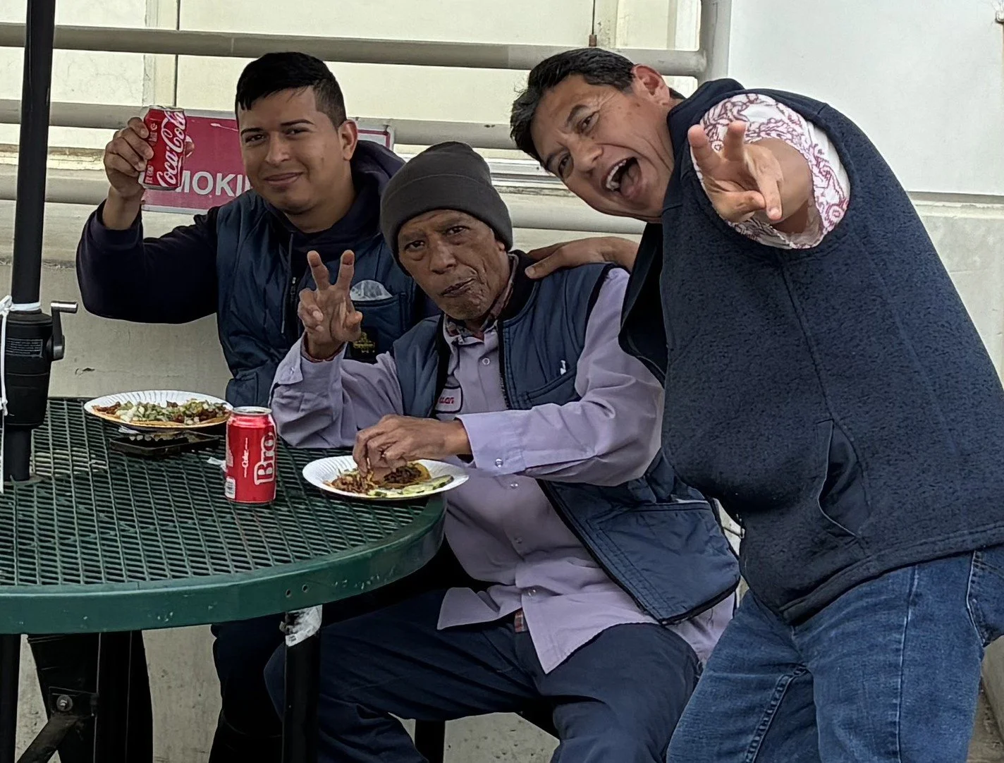 Three men outdoors at a green metal table with food and drinks, posing and smiling for the camera.