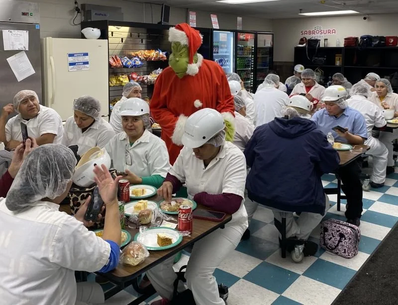 People in a cafeteria wearing hairnets and uniforms, with a person dressed as the Grinch in a Santa hat and red outfit, celebrating or distributing food.