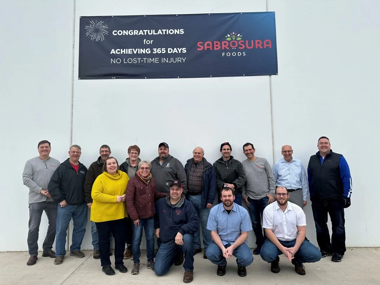 Group of 14 people standing and squatting in front of a white wall with a banner that reads "Congratulations for achieving 365 days no lost-time injury" and a logo for Sabrosura Foods. The group appears to be celebrating an achievement.