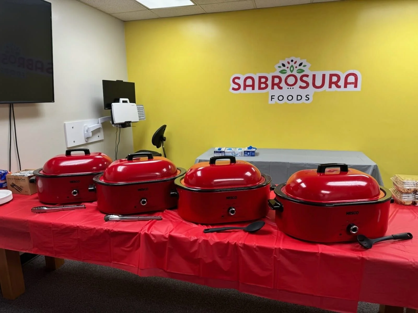 Four red slow cookers lined up on a table with a red tablecloth, set up for a buffet at Sabrosura Foods. There are serving utensils in front of each slow cooker. In the background, a yellow wall features the Sabrosura Foods logo, and there are some b