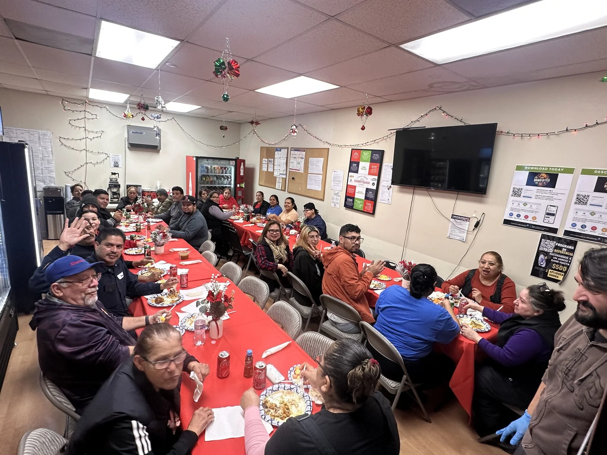 A group of people gathered around long tables with red tablecloths, celebrating a holiday or special occasion in a decorated room with Christmas ornaments hanging from the ceiling.