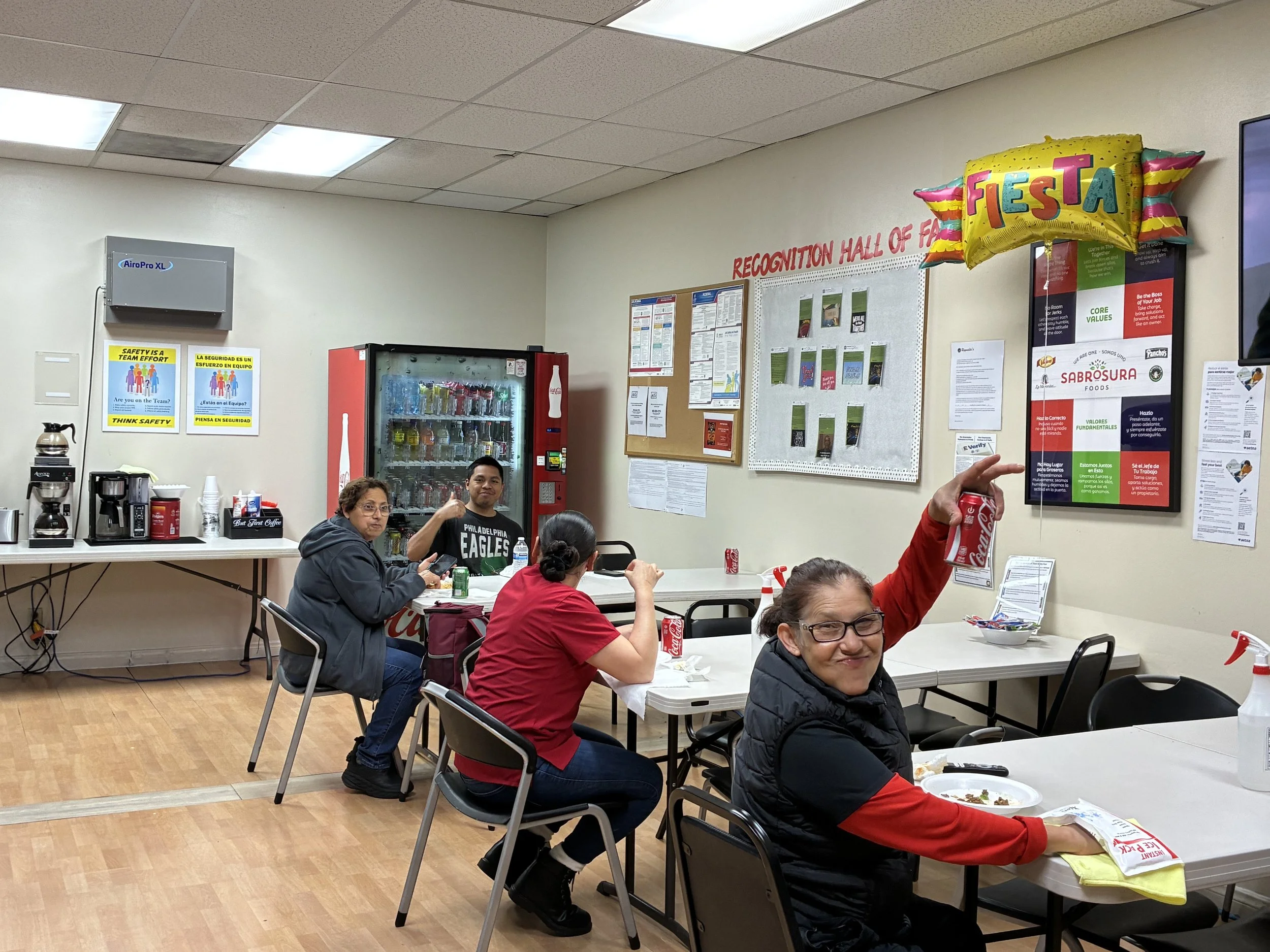 Four people sitting at tables in a break room, three women and one man, with snacks and drinks, smiling and gesturing. The room has a vending machine, signs, and a colorful fiesta balloon decoration.