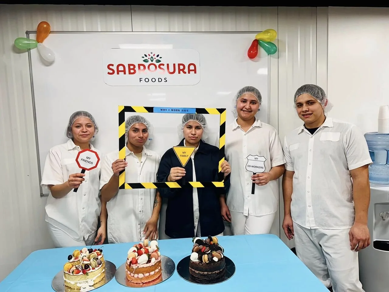 Five women in white uniforms and hairnets standing behind a table with three decorated cakes. They hold signs reading "My Friends," "My Family," and "My Co-Workers." Behind them is a whiteboard with the logo "Sabrosura Foods" and colorful balloons.