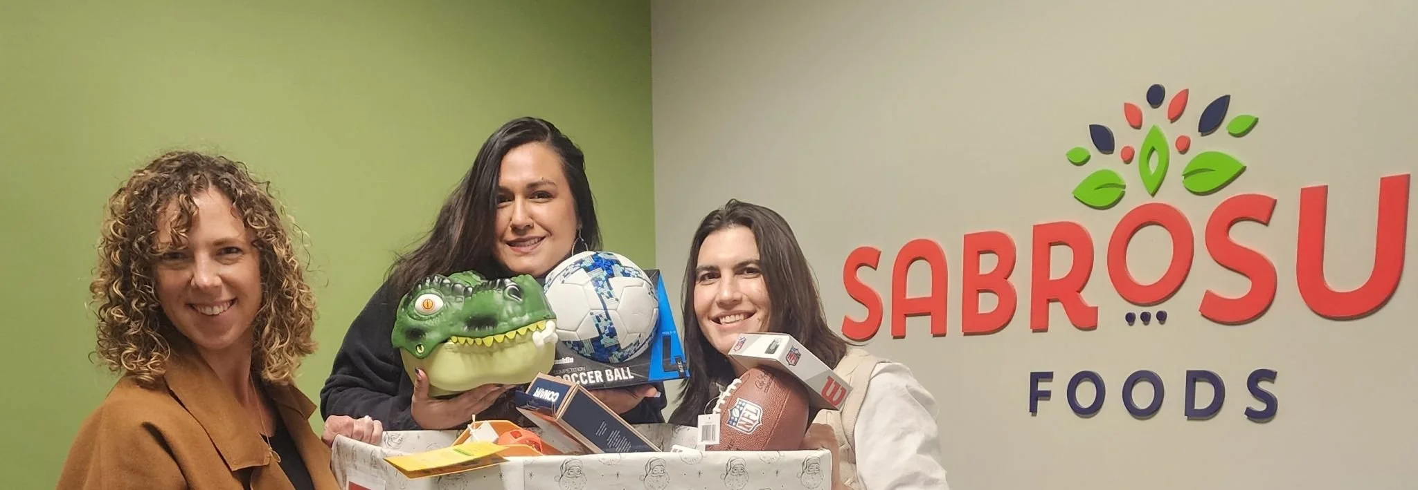 Three women smiling and holding toys and a gift basket inside Sabroso Foods, with a green and beige wall background, and the Sabroso Foods logo visible.