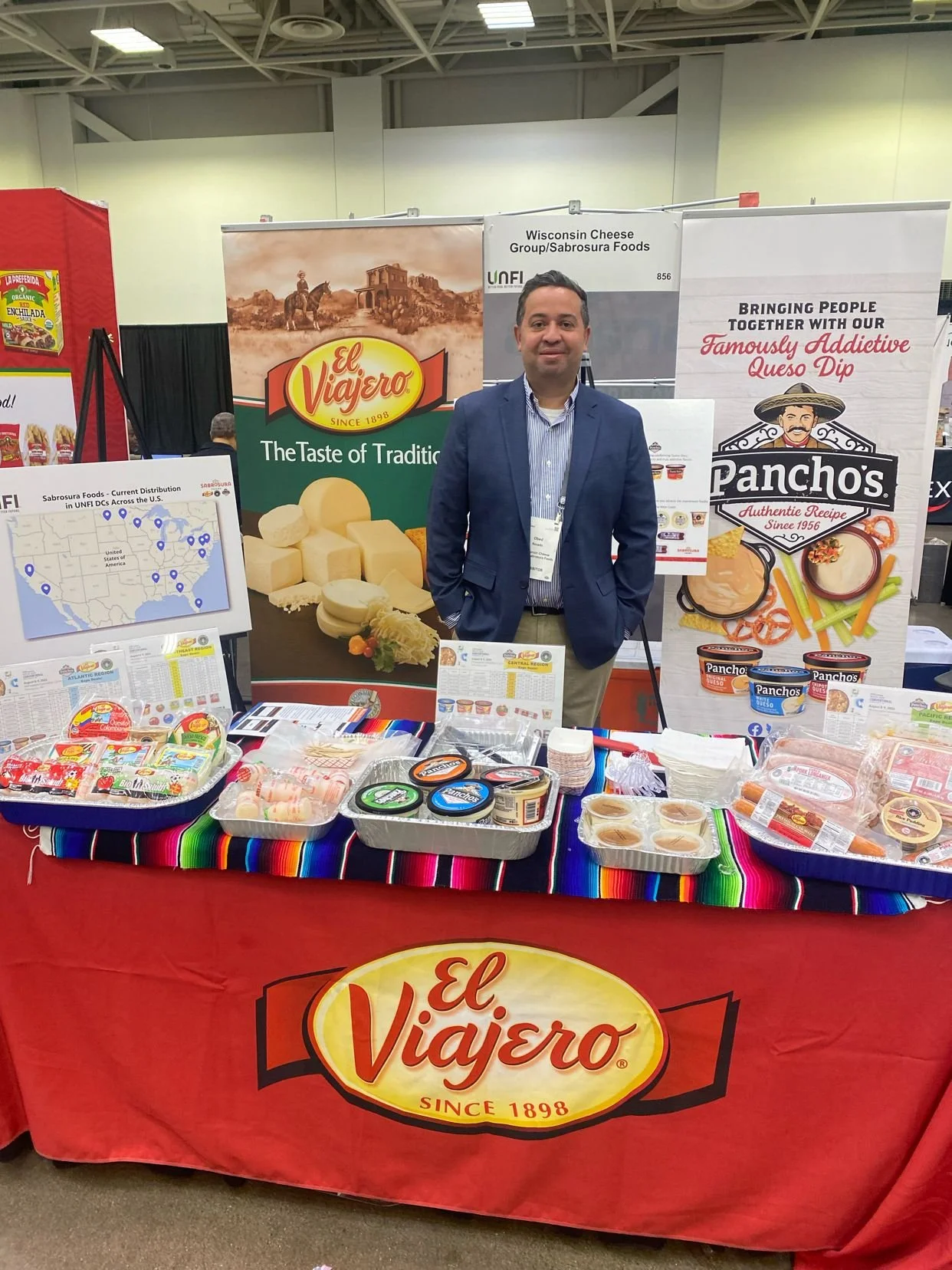 A man standing behind a booth displaying El Viajero cheese and Pancho's queso dip at a trade show. The booth has banners, product samples, and a colorful tablecloth with Mexican cultural colors.