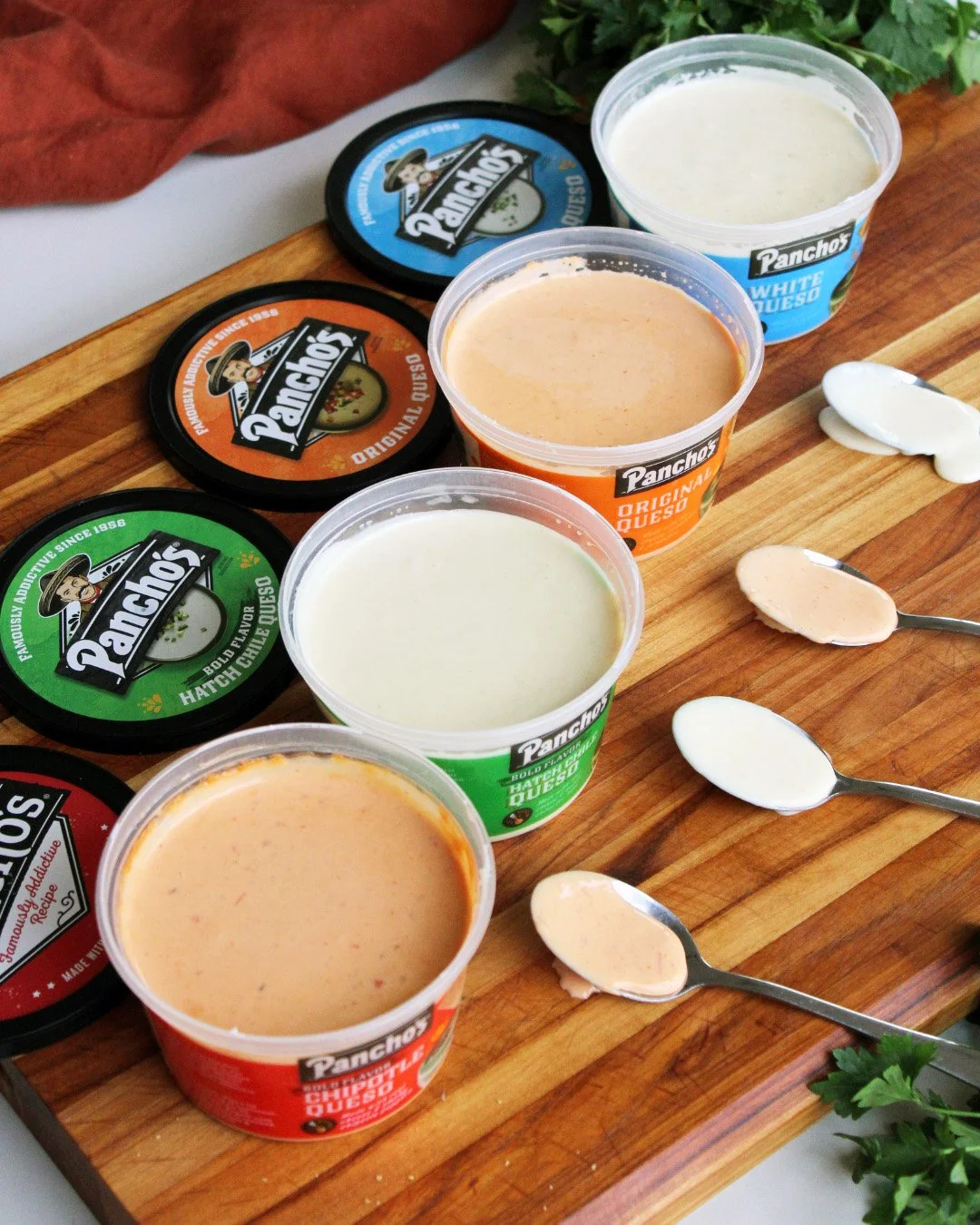 Assortment of five cups of Pancho's queso on a wooden tray, with small spoons of different queso flavors and cilantro garnish in the background.