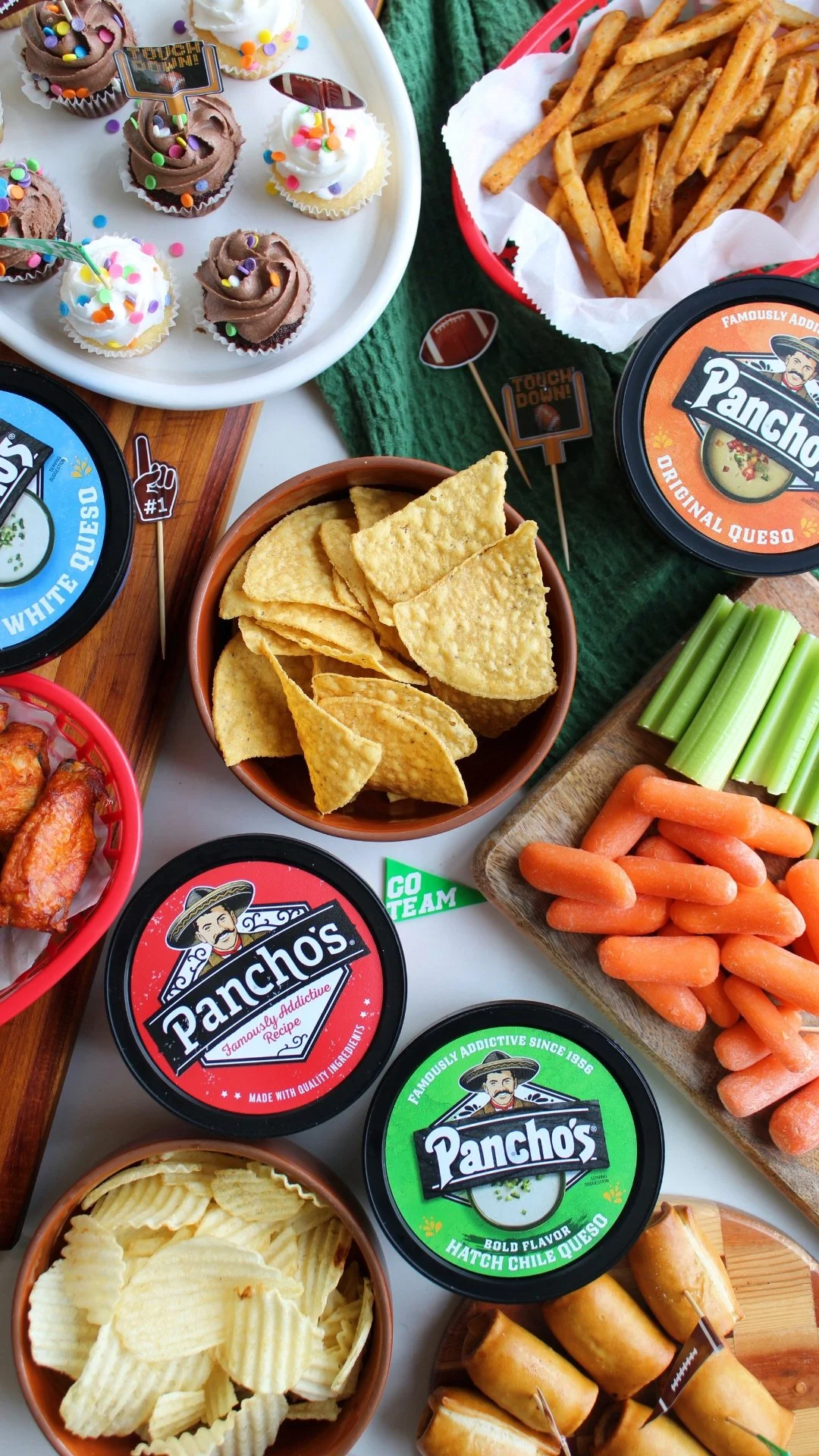 A variety of snack foods including mini cupcakes with colorful sprinkles, French fries, tortilla chips, carrot sticks, celery sticks, potato chips, chicken wings, and dips from Pancho's brand on a wooden and white table with green and red accents.