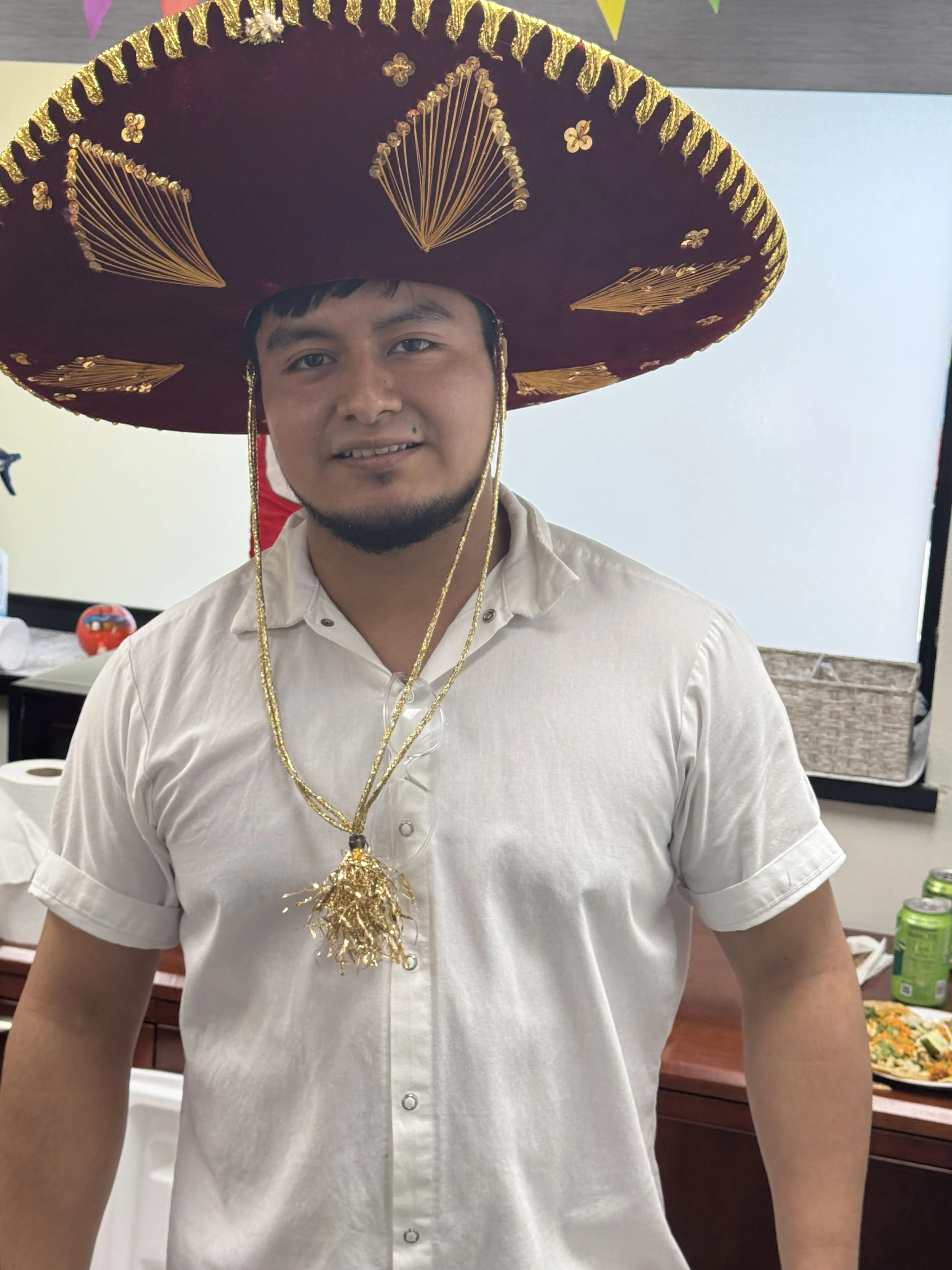 A man wearing a large maroon and gold sombrero with gold decorations, a beige short-sleeved button-up shirt, and gold tinsel necklaces, standing indoors with a decorated background.