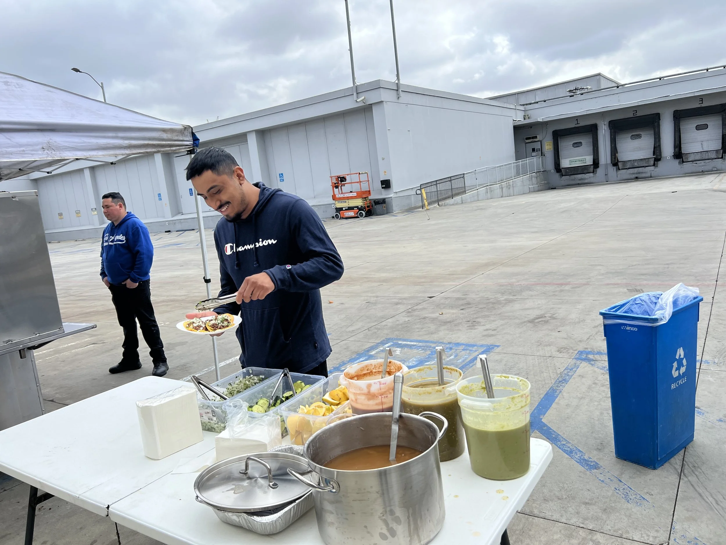 A young man serving food from a table with tubs of salsa and sauces at an outdoor event, with another person standing in the background, set up in a parking lot outside a warehouse or distribution center.