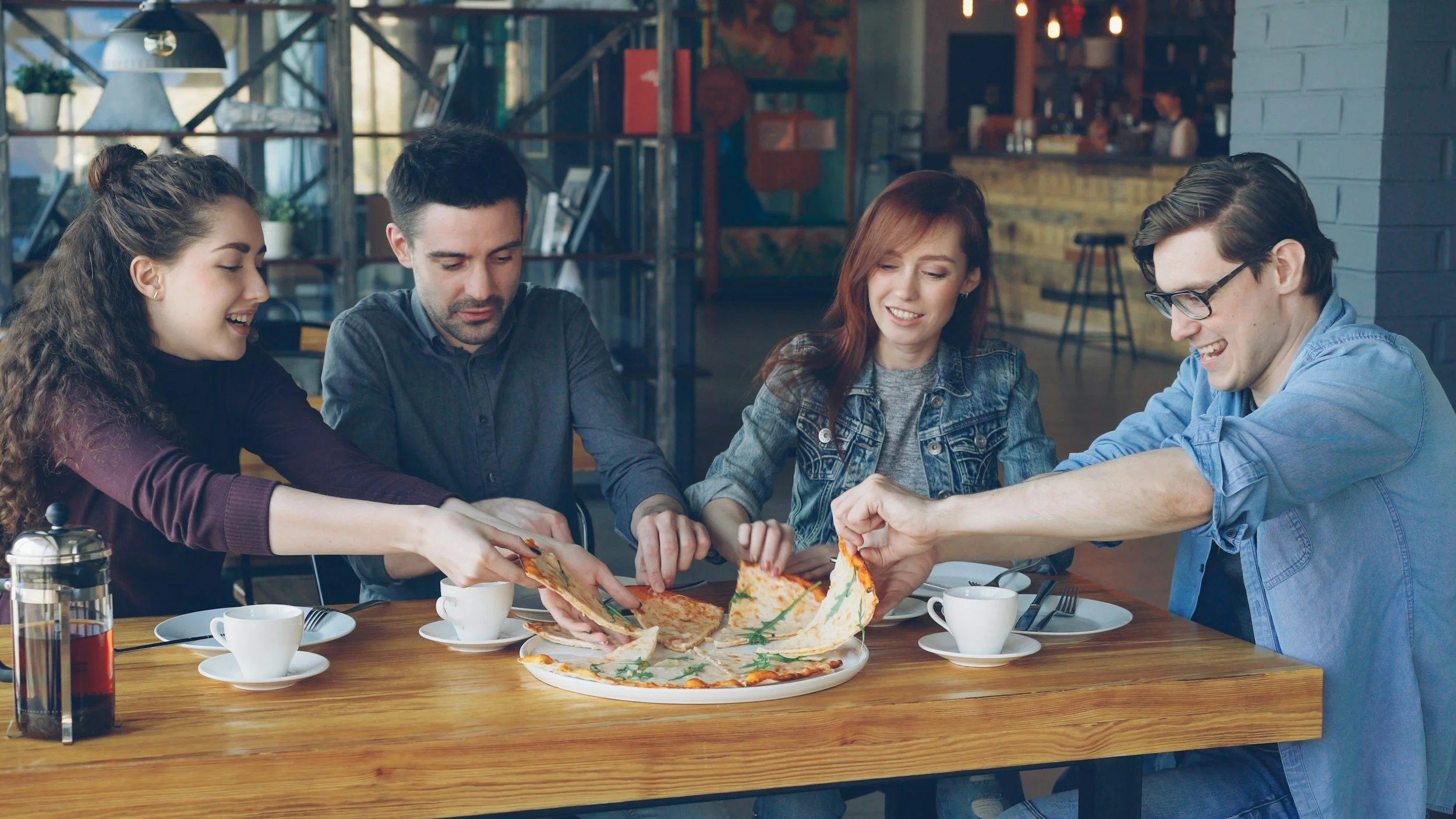 Four people sitting at a table reaching for slices of pizza together.