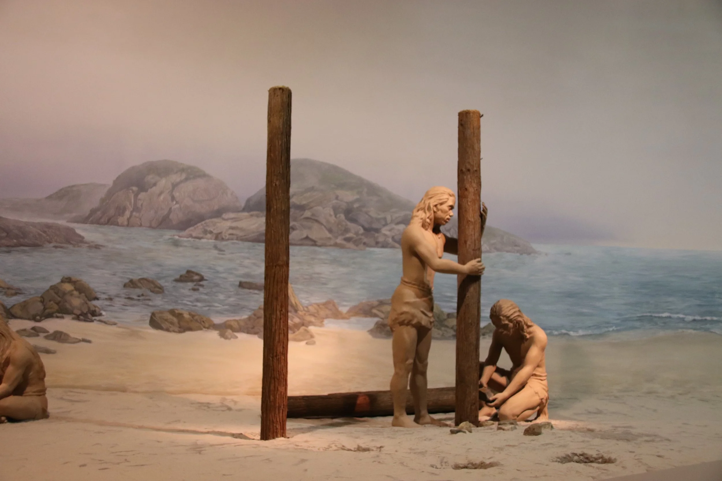 Neolithic human figures building a wooden structure on a sandy Hong Kong shoreline in a museum diorama, with rocky coastal scenery and ocean in the background.