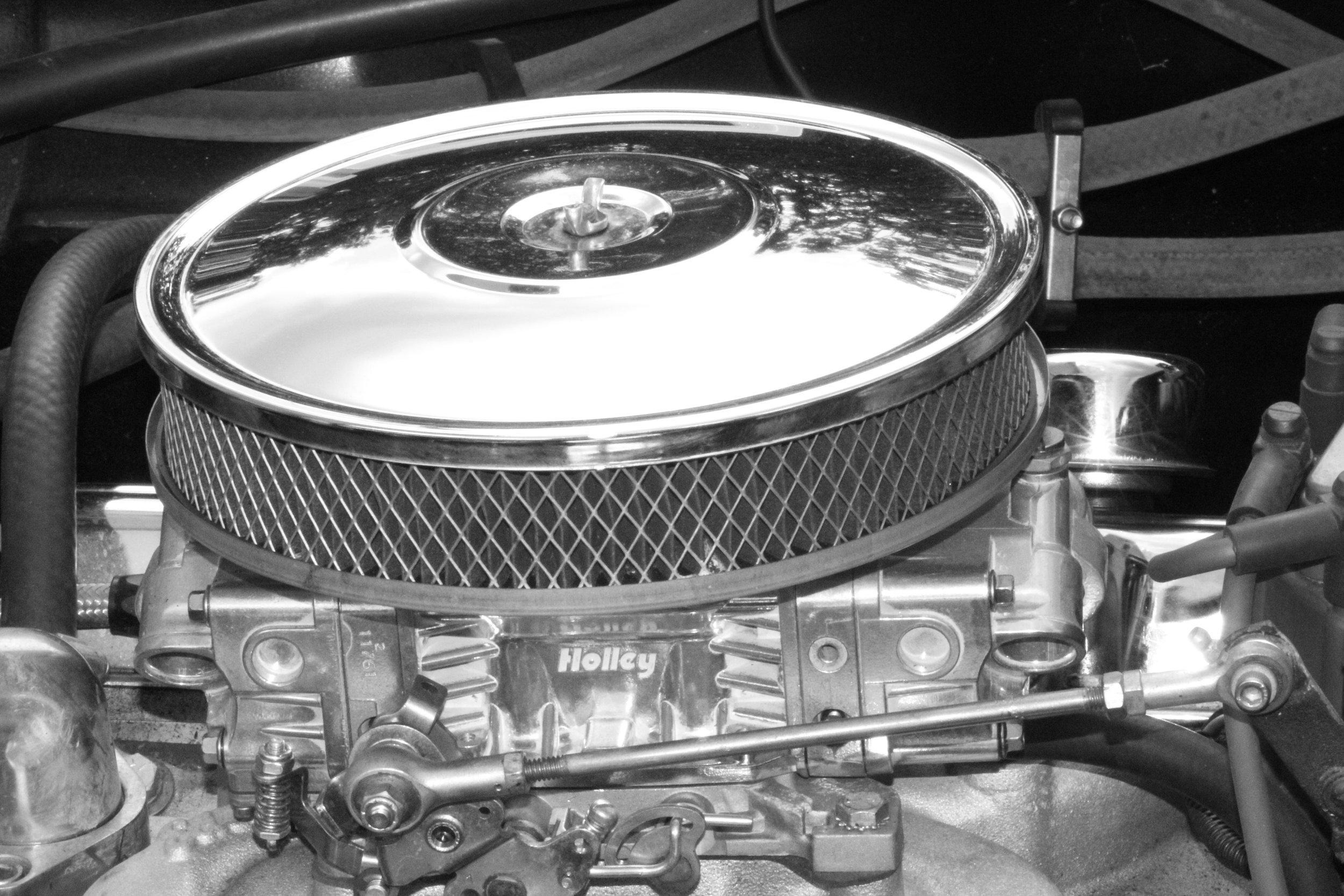 Black and white photo of a car engine with a chrome air filter on top and various mechanical parts and hoses surrounding it.