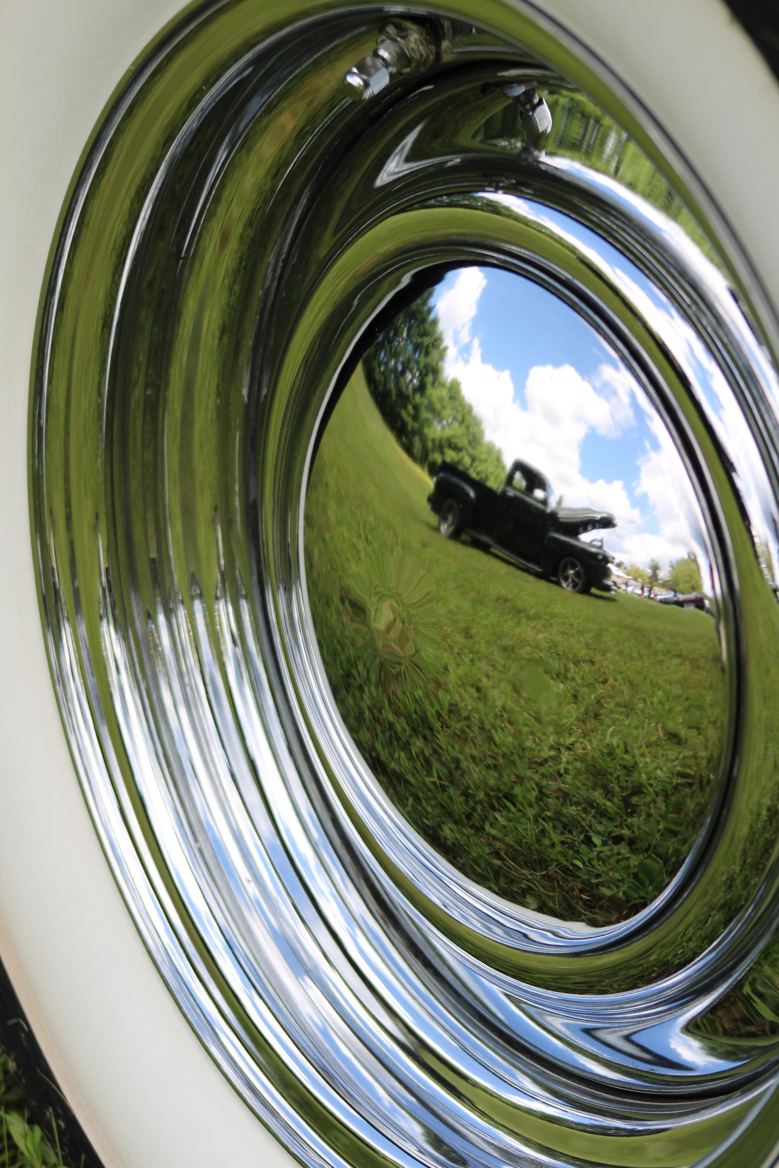 Reflection of a black pickup truck and trees on a shiny, circular, chrome surface of a washing machine drum outdoors under a partly cloudy sky.