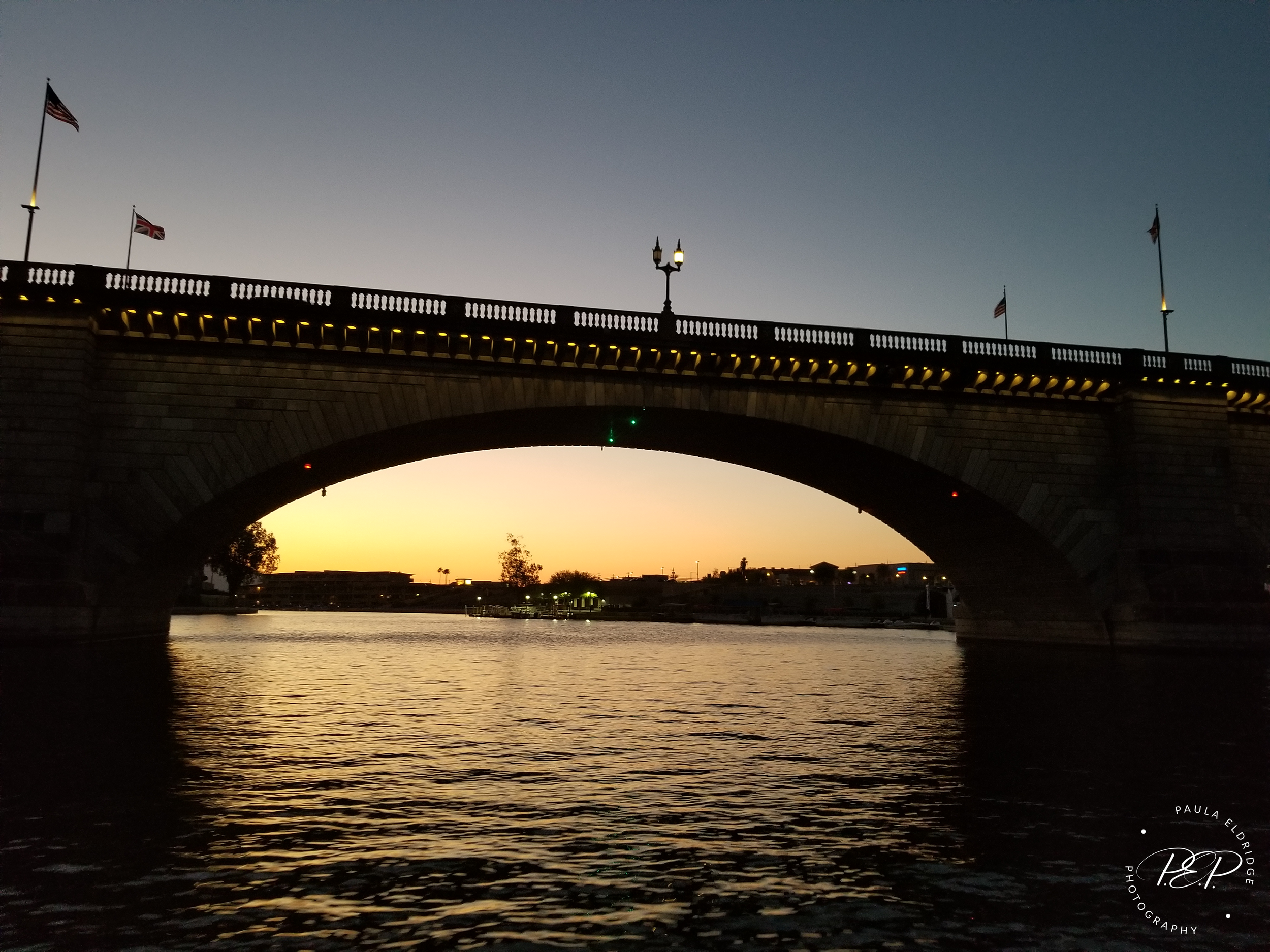 A bridge over a body of water at sunset, with flags on top of the bridge and lights along the bridge's edge.