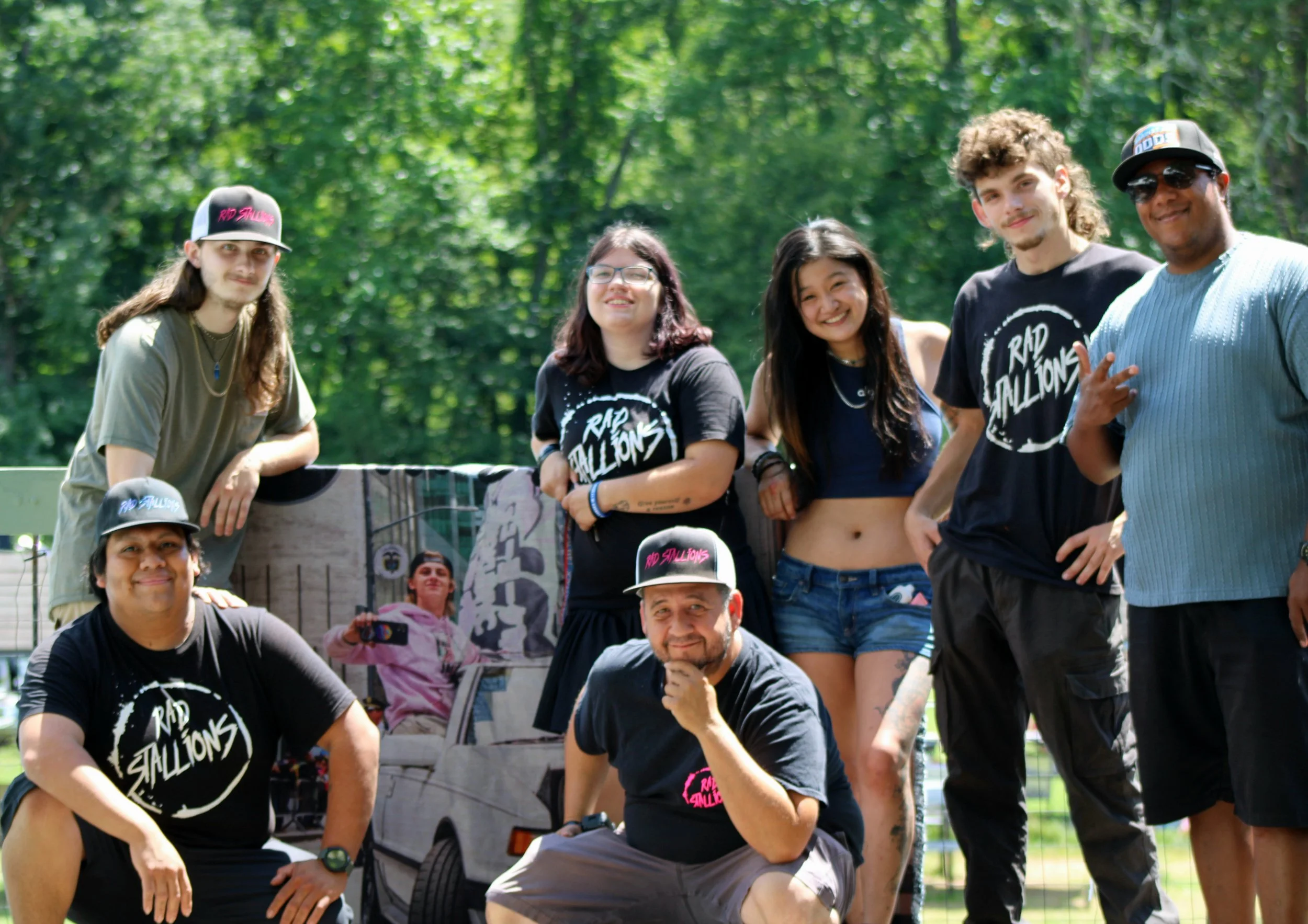 A group of six smiling friends posing outdoors on a sunny day, standing in front of a promotional cart for the band 'Rad Stallions', with trees and greenery in the background.