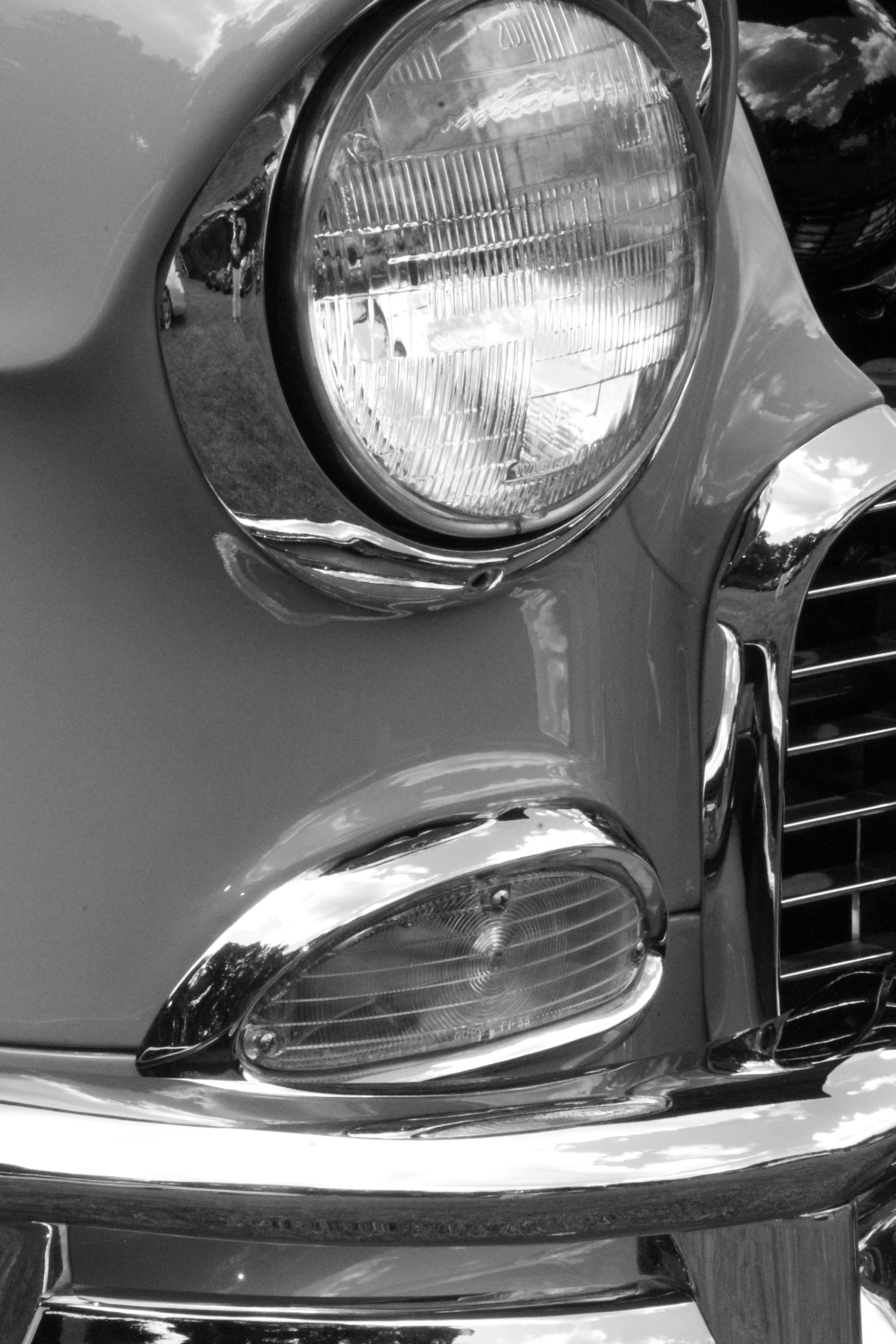 Close-up of a car's front headlight and bumper, with a reflection of the sky and trees in the car's shiny surface, in black and white.