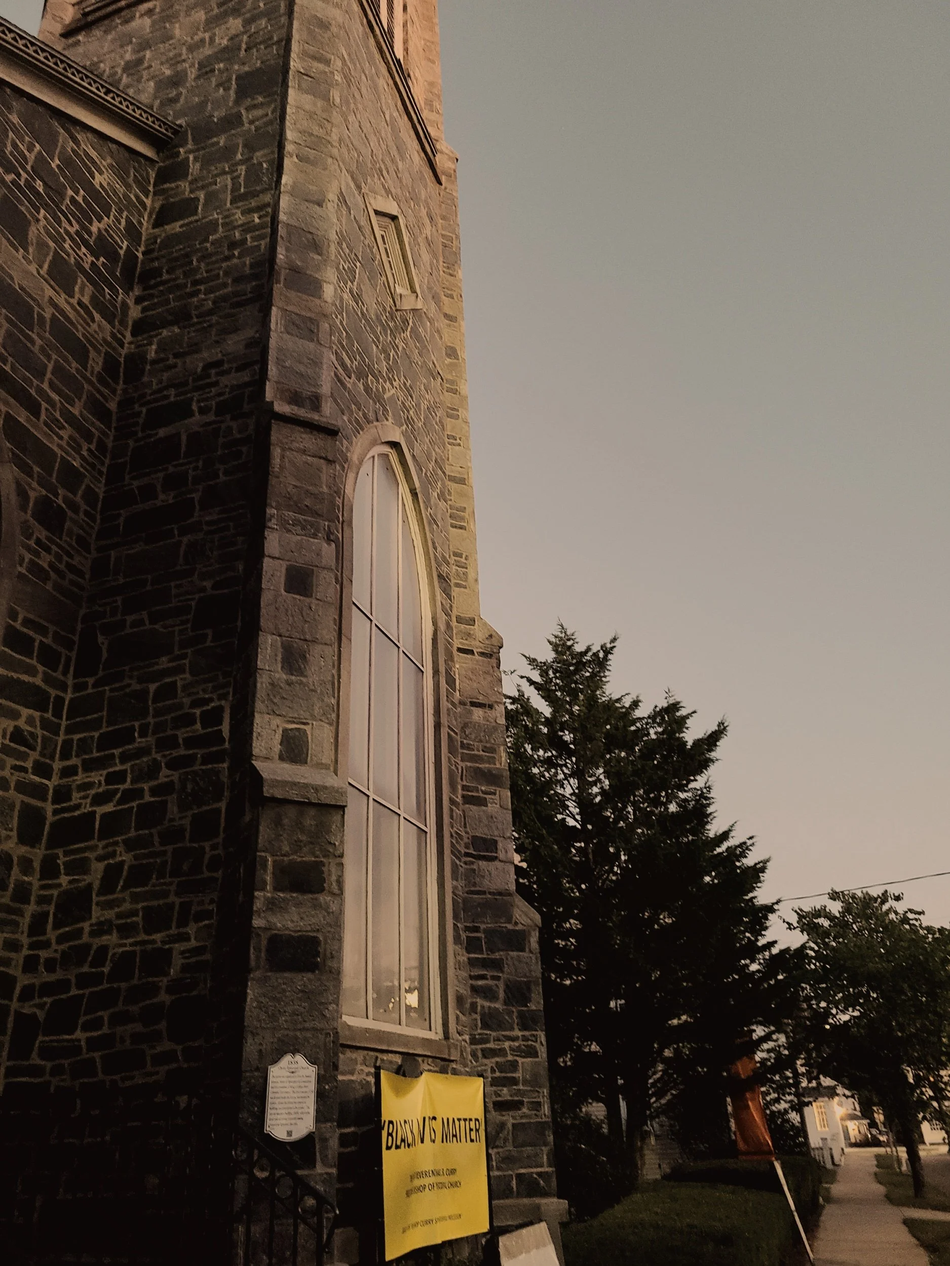 A stone church or building with a tall arched window. There is a yellow sign that reads 'Black Lives Matter' on a black iron fence. Trees and a street are visible in the background.