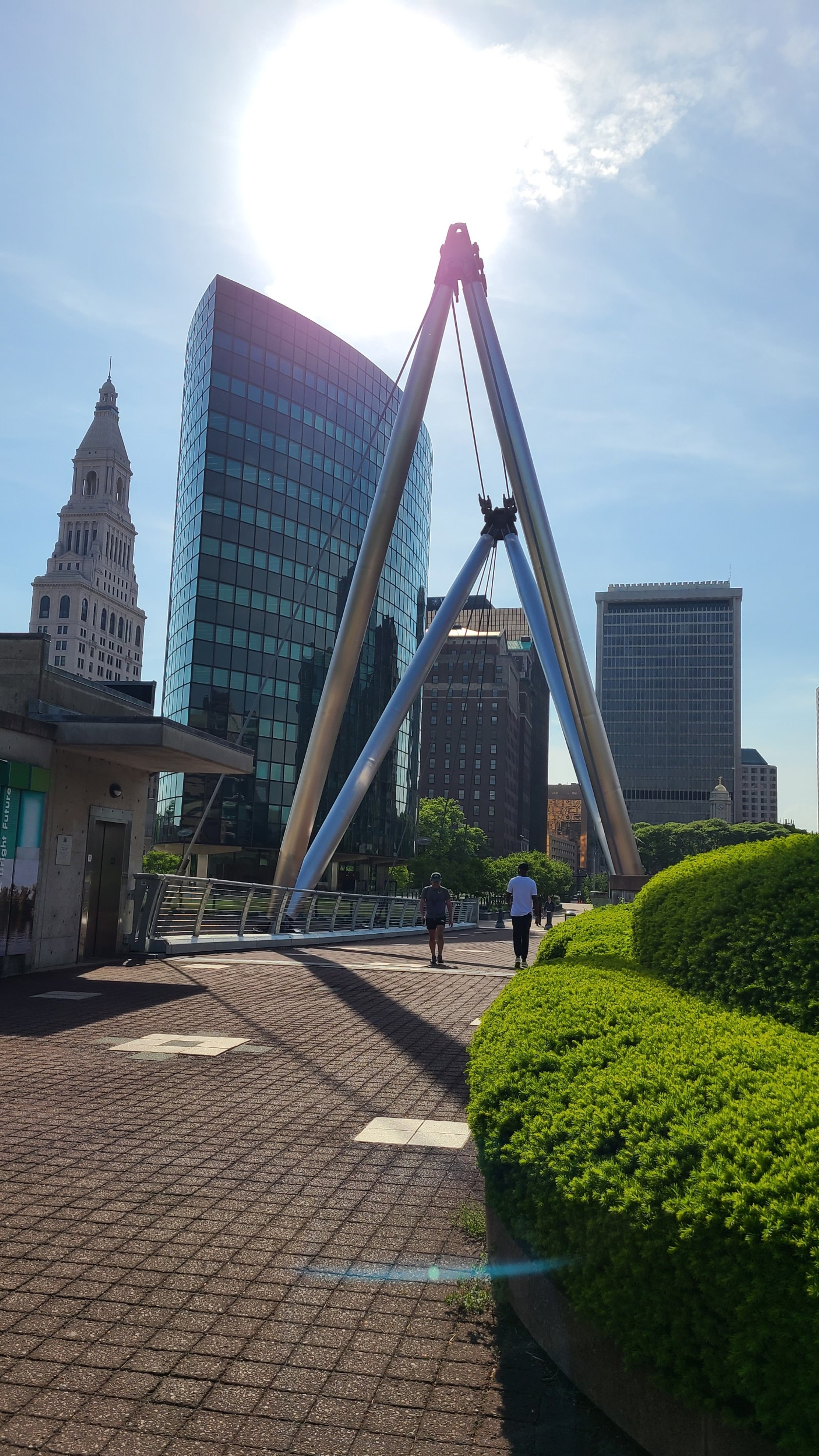 Urban park with modern glass buildings, a tall tower, and people walking on a paved path with greenery and bushes.