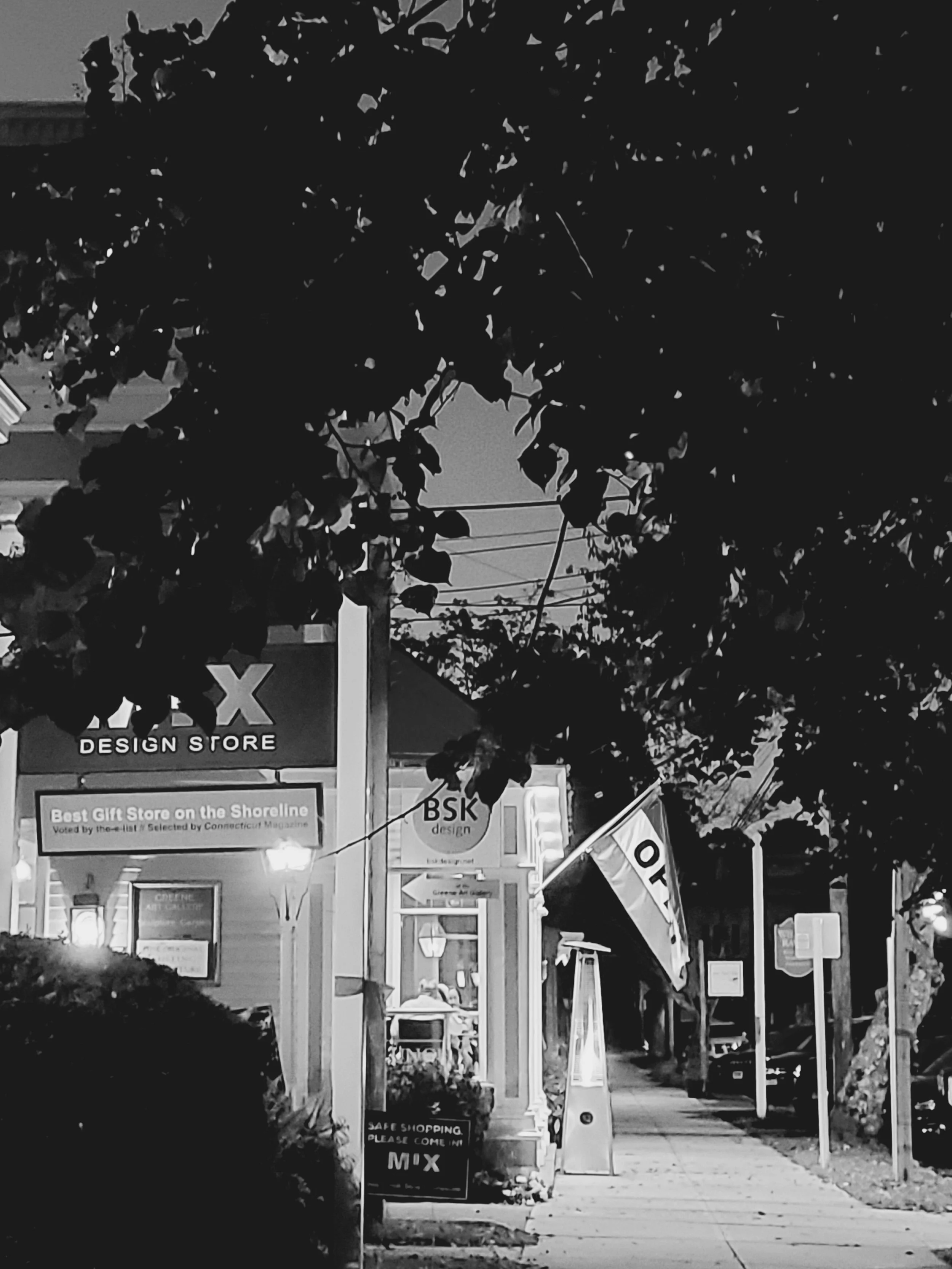 A sidewalk scene at dusk with a store sign that reads 'MIX DESIGN STORE' and another sign that says 'Best Gift Store on the Shoreline.' There are trees, streetlights, and a few objects including a flag and outdoor heaters. The scene is in black and w