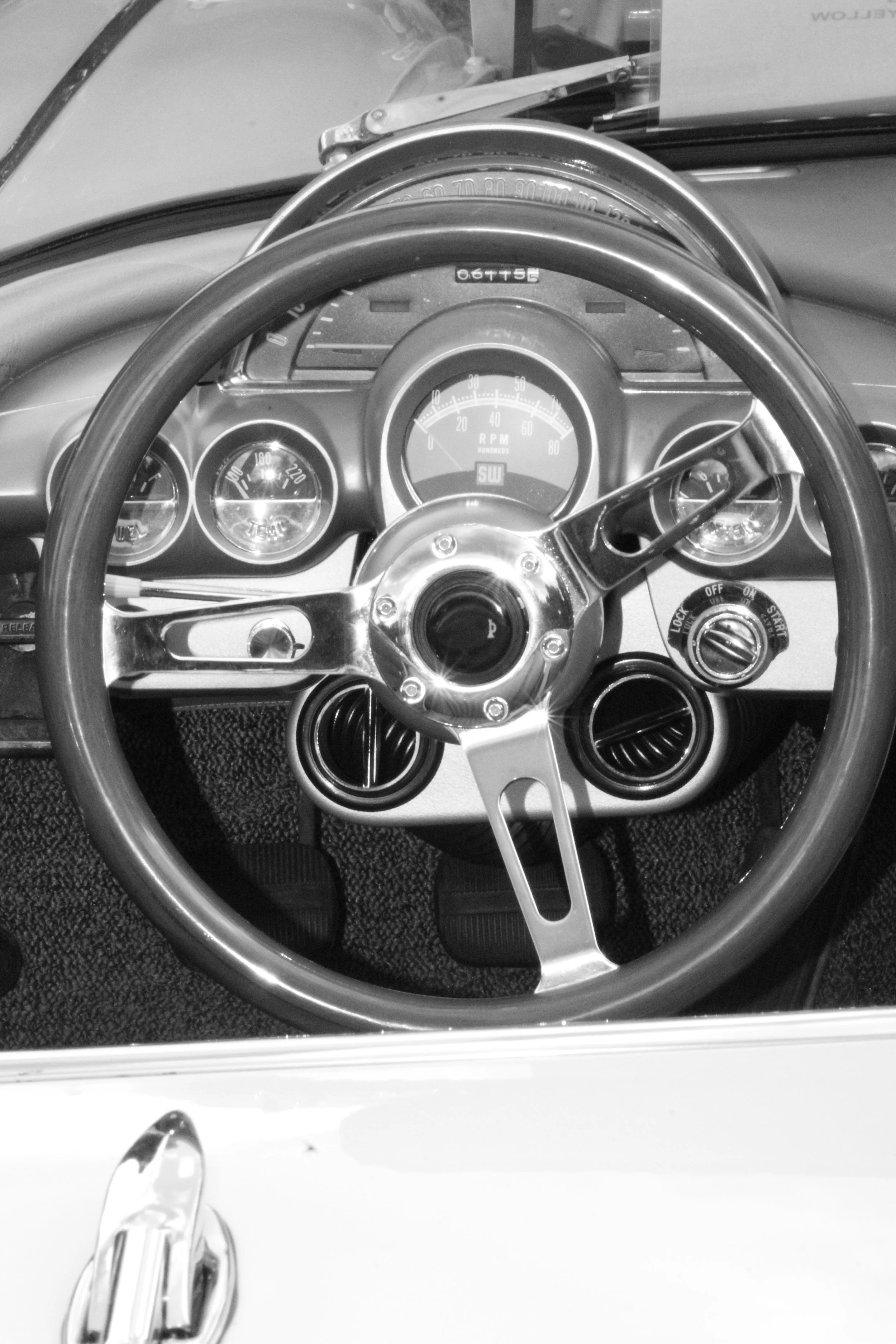 Black and white photo of a vintage car dashboard showing a steering wheel, speedometer, tachometer, and other gauges, with a partially visible dashboard and side mirror.