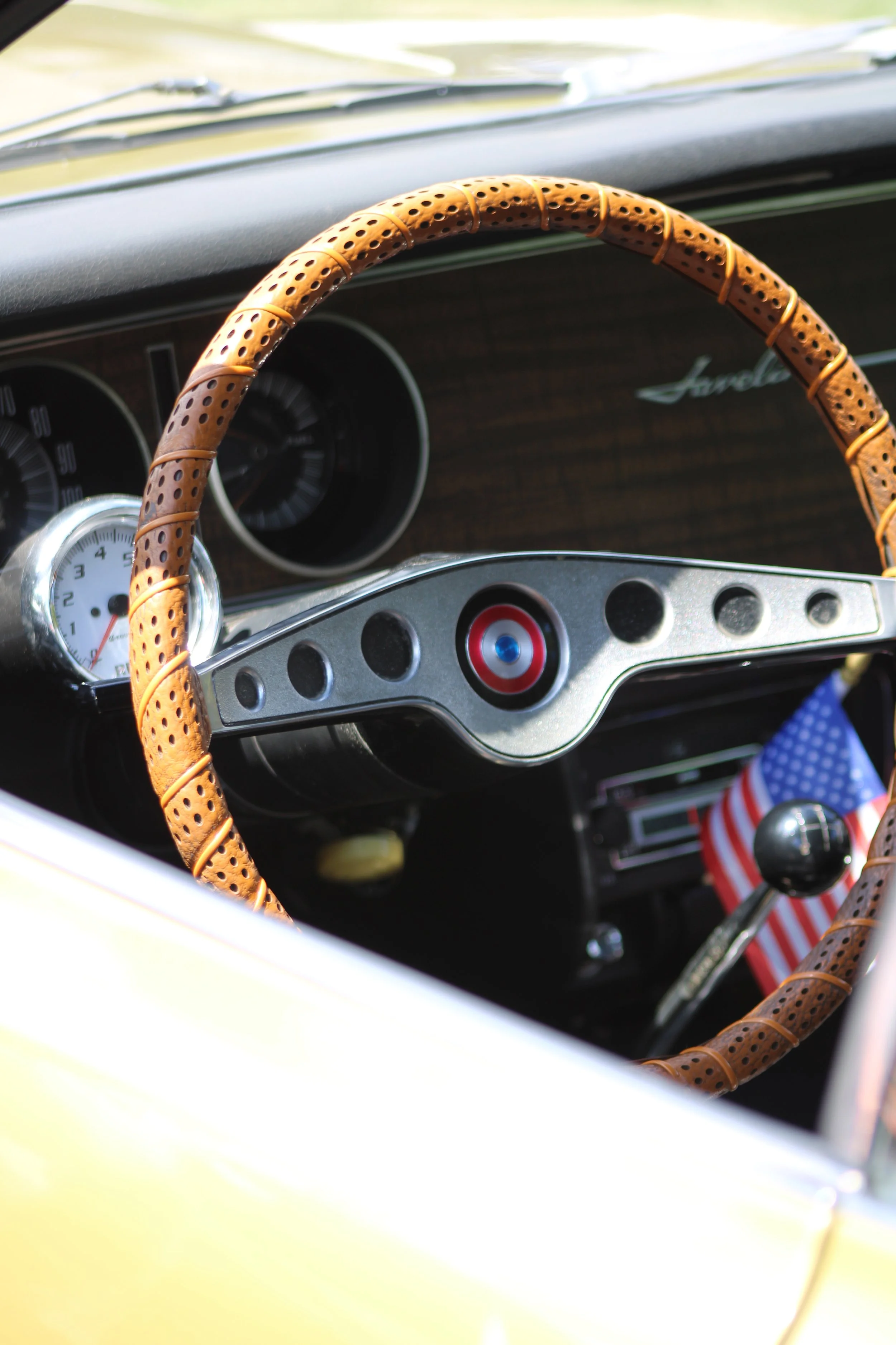 Close-up of a vintage car dashboard with a perforated leather steering wheel cover, speedometer, and Garrard logo, with American flag-themed fabric on the gear shifter.