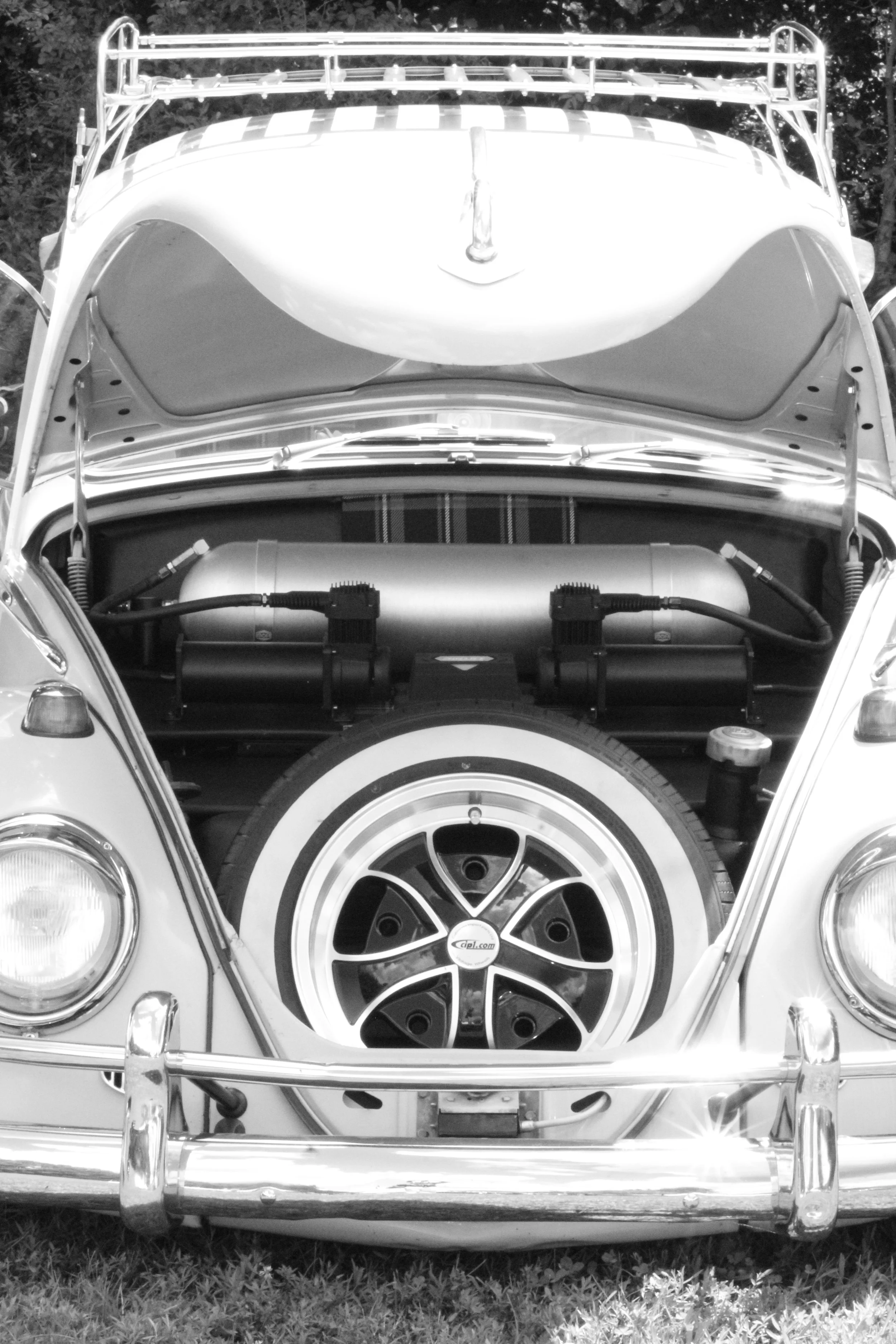 Front view of a classic car with its hood open, showing the engine. The car has a round headlight, a distinctive wheel with a star-like pattern, and a chrome bumper. The car is parked on grass, and trees are visible in the background.