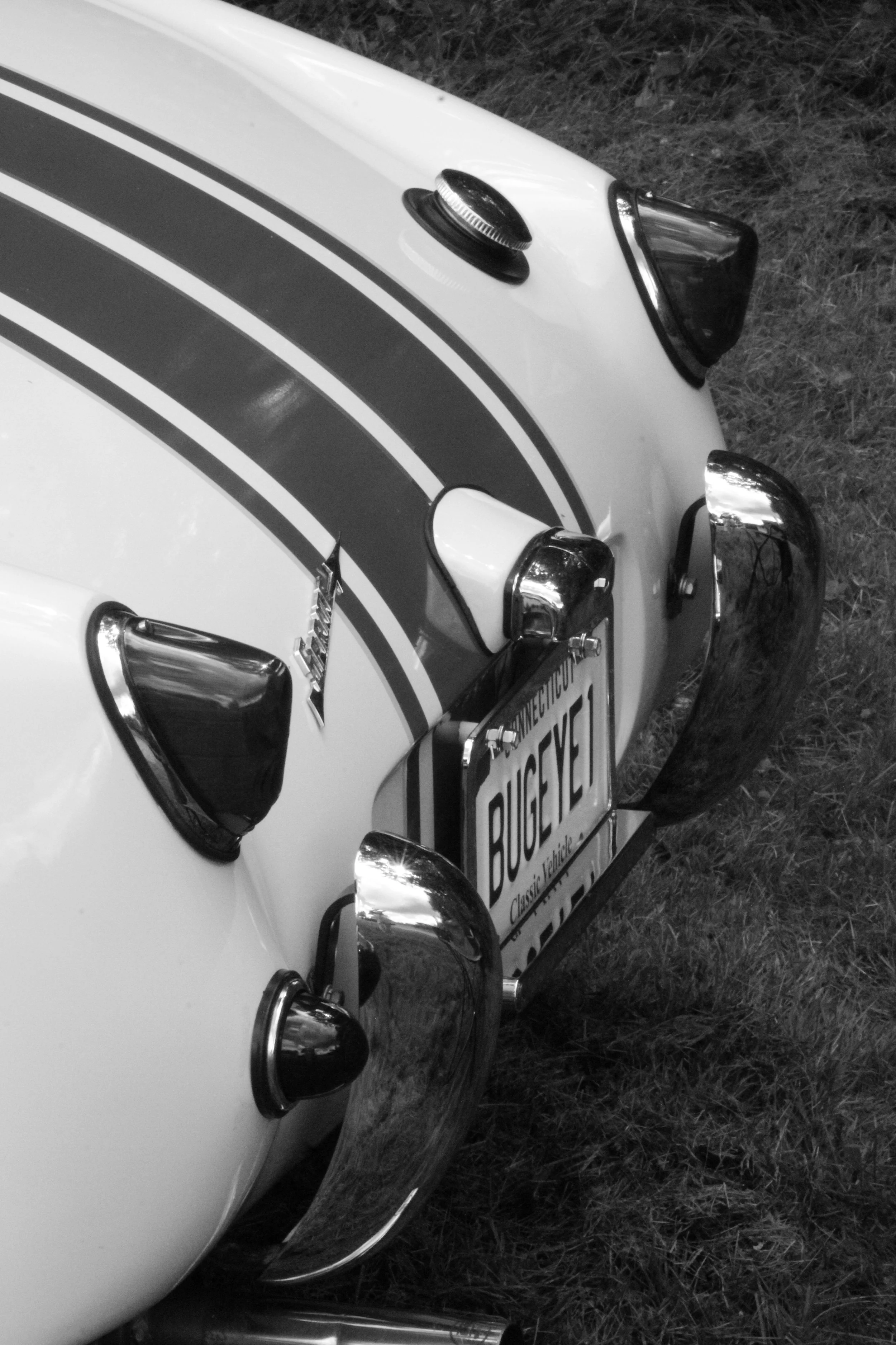 Close-up of the front of a vintage white sports car with racing stripes, showing headlight, side mirror, hood ornament, and a Connecticut license plate reading 'BUGSY'.