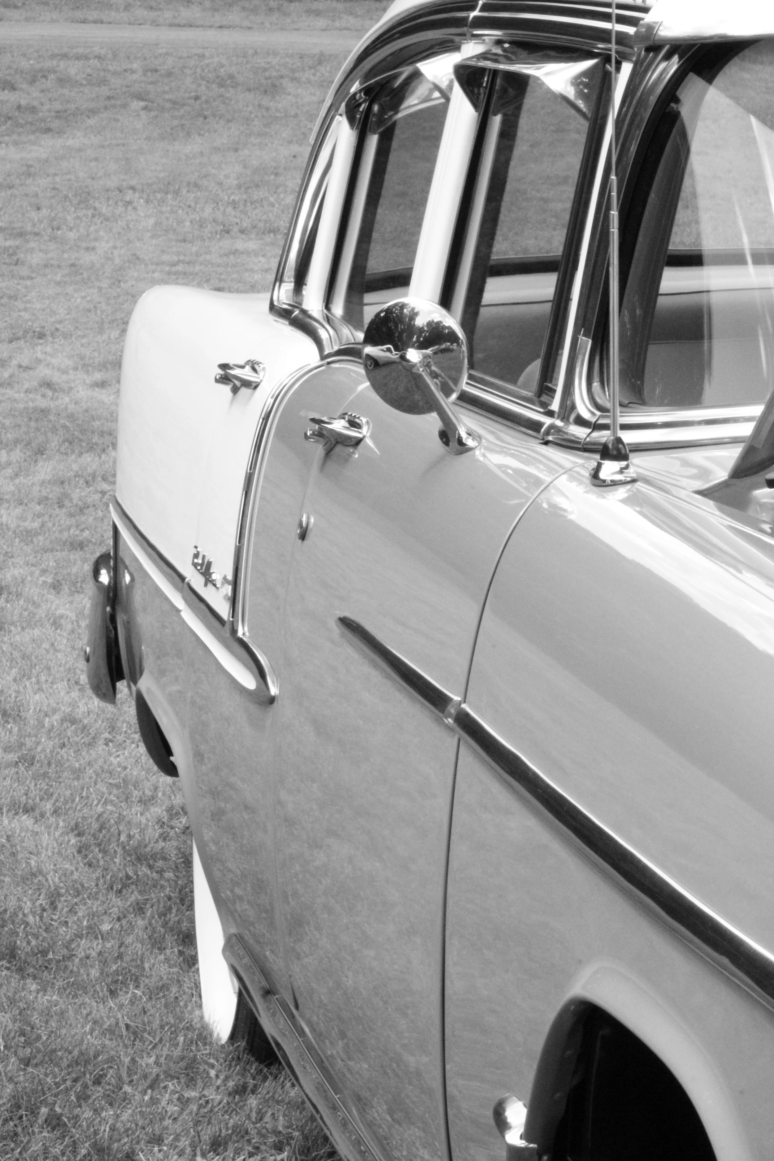Black and white photo of a vintage car parked on grass, showing the front side including the side mirror, door handle, window, and part of the wheel.