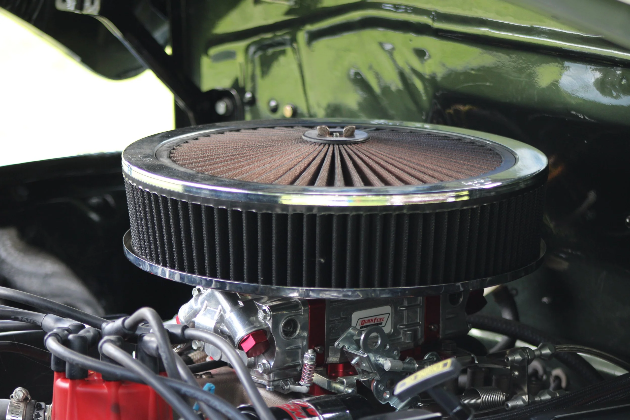 Close-up of a car engine with a large round air filter in the center, surrounded by various engine components and wiring, with a green painted engine bay in the background.