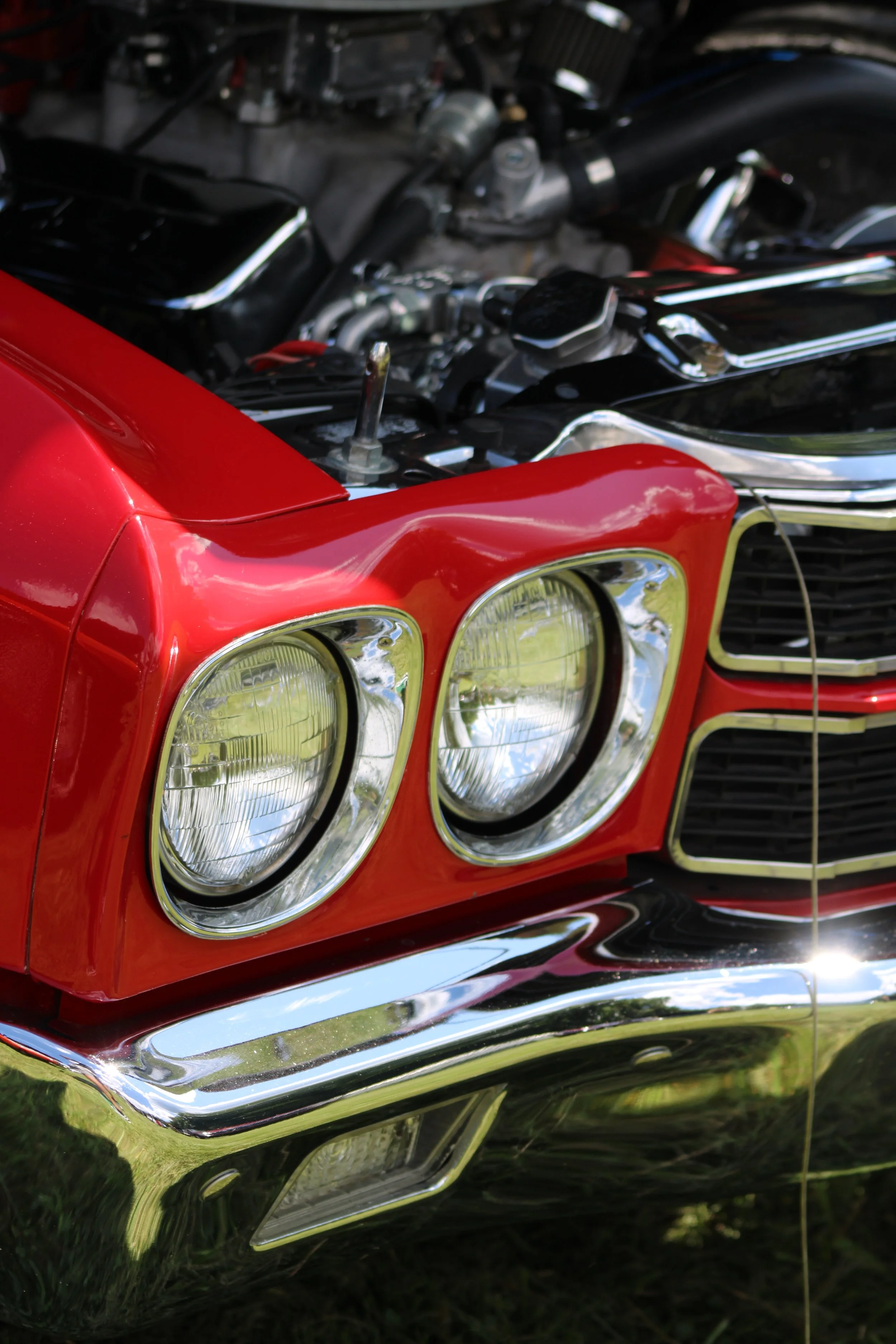 Close-up of a classic red car's front with dual headlights and chrome bumper, showing a detailed view of the car's grille and hood.