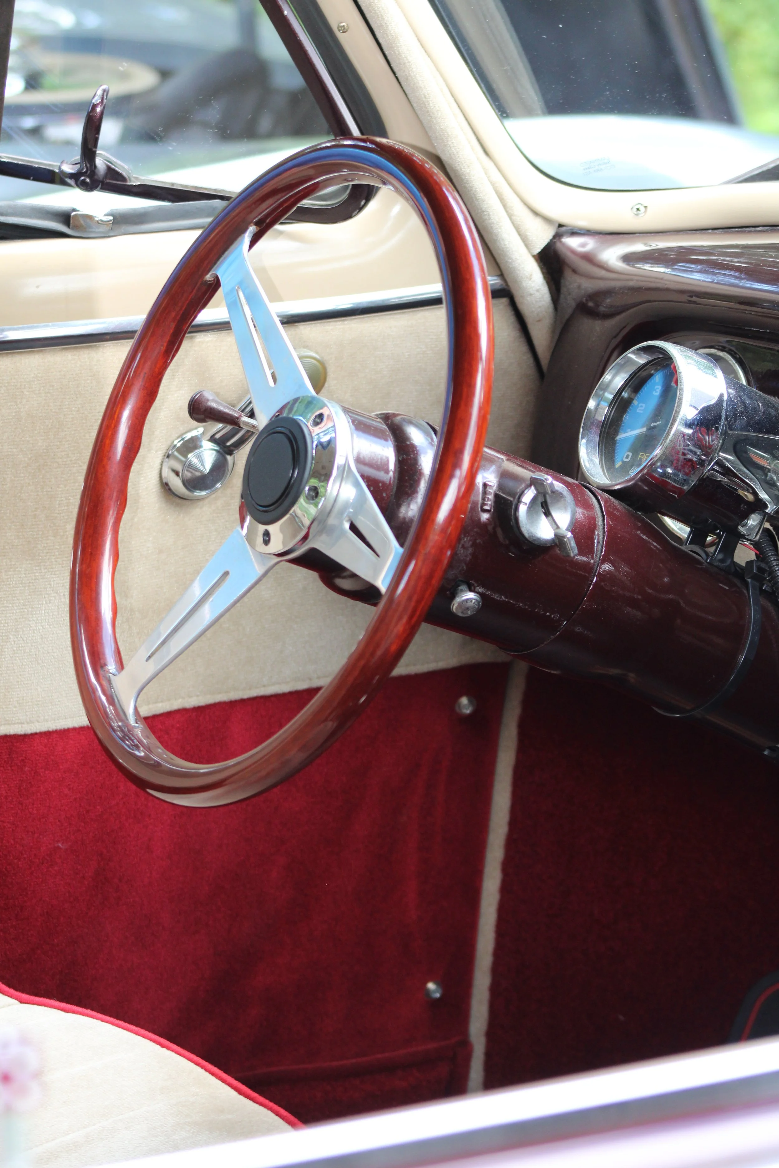 Interior of a vintage car, focusing on the steering wheel with a wood finish and chrome spokes, and a gauge cluster on the dashboard.