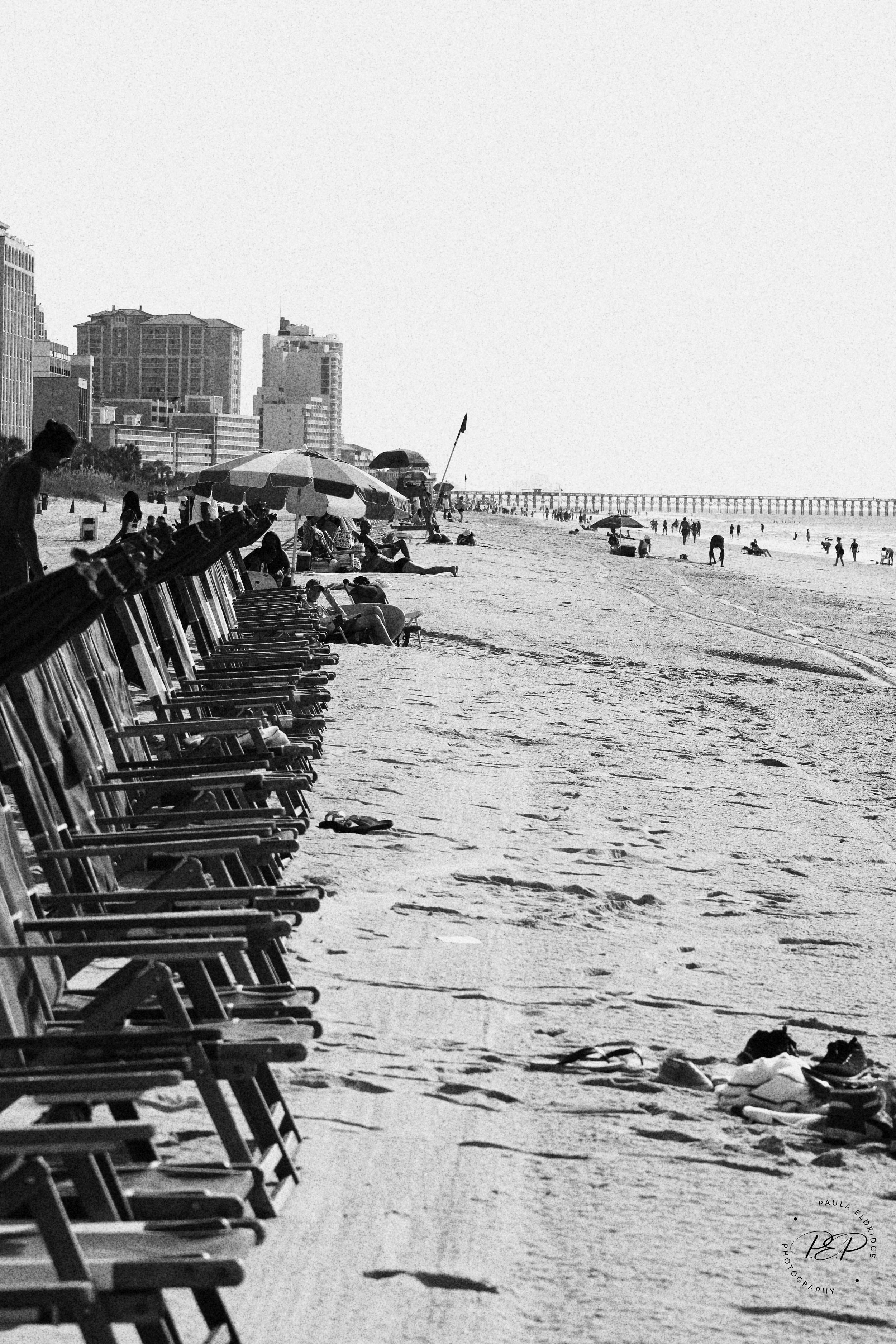Black and white image of a crowded beach with lounge chairs and umbrellas, along a shoreline with people walking and relaxing, city buildings in the background.