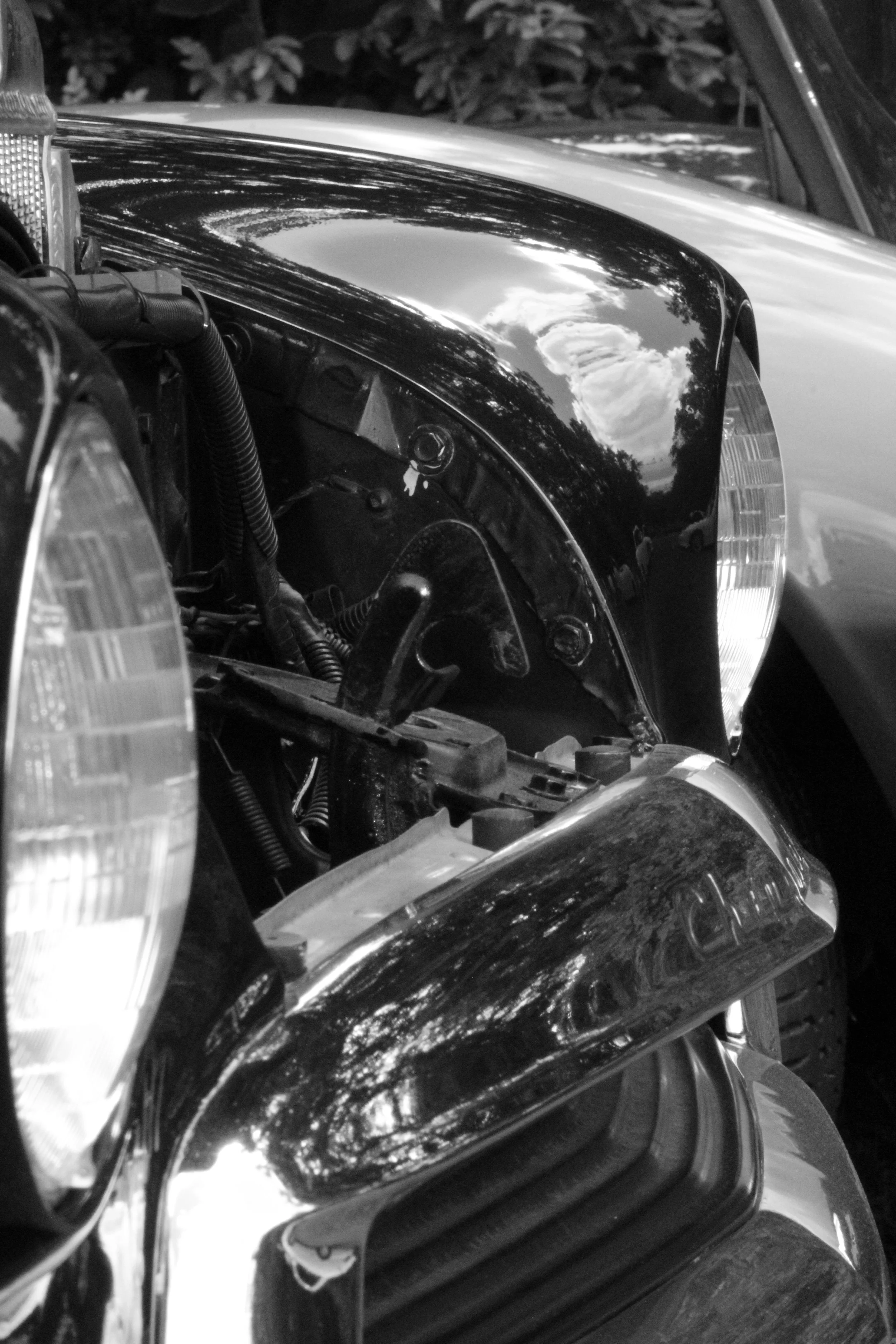 Close-up of a motorcycle's front section showing its headlight, reflection of the sky and trees on the shiny black surface, and parts of the engine.