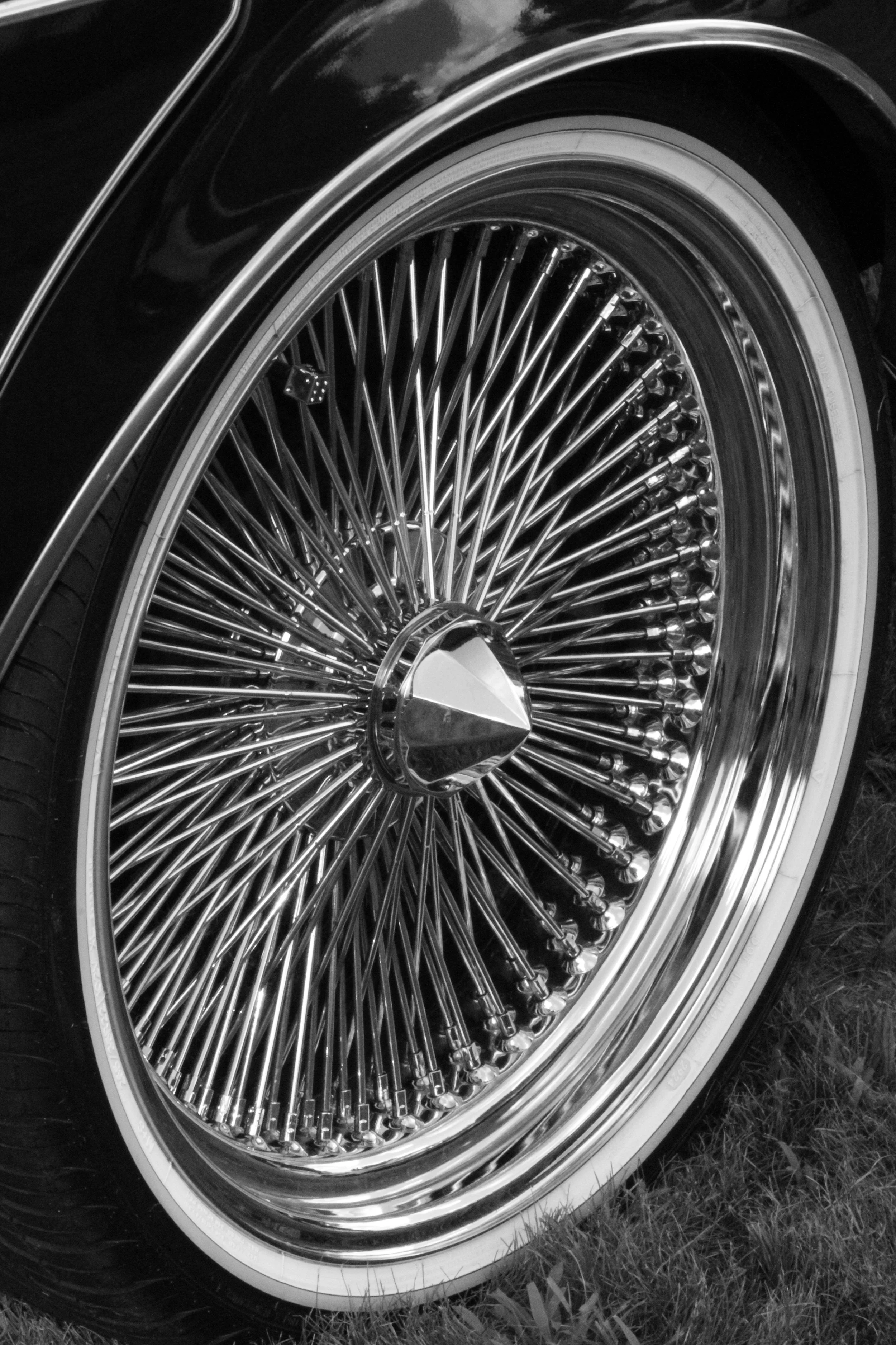 A close-up black and white photo of a vintage car wheel with a wire-spoke rim and a tire, resting on grass.