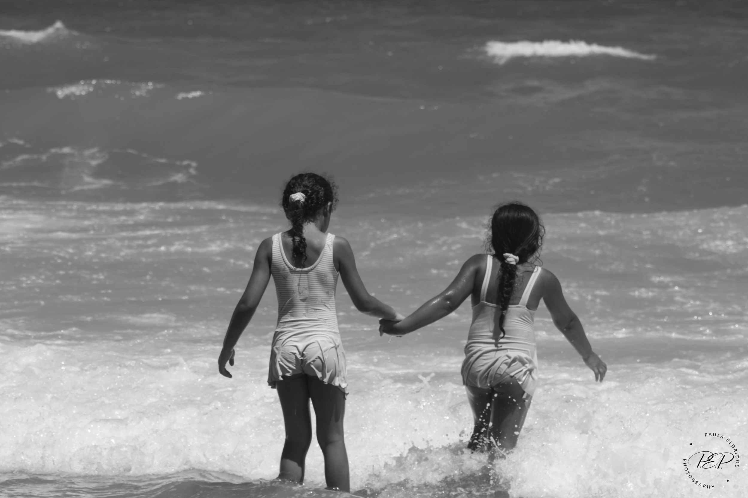 Two young girls holding hands and walking into the ocean waves at the beach.