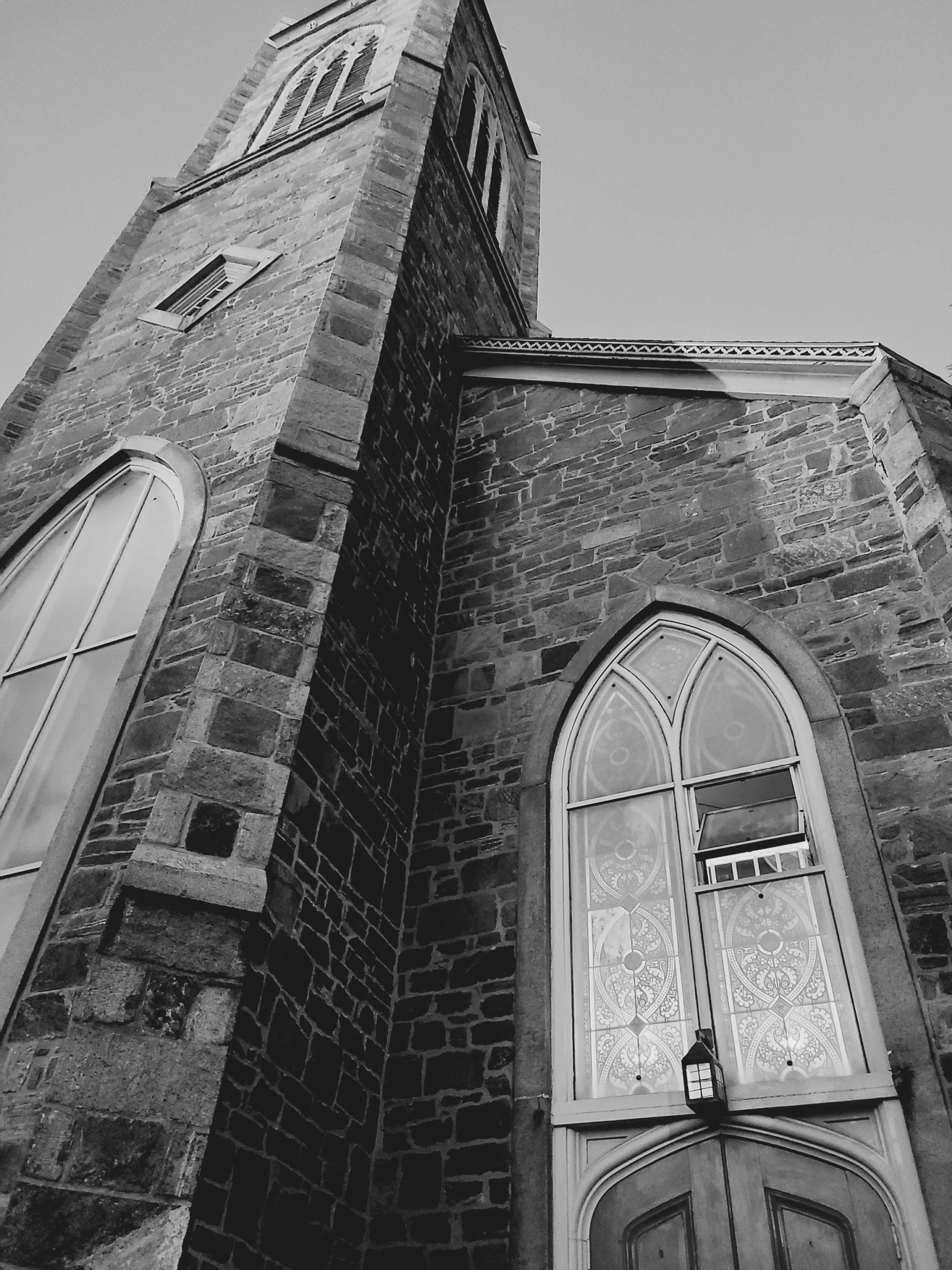 A black and white photo of a tall stone church or cathedral with pointed arched stained glass windows and a clock tower.