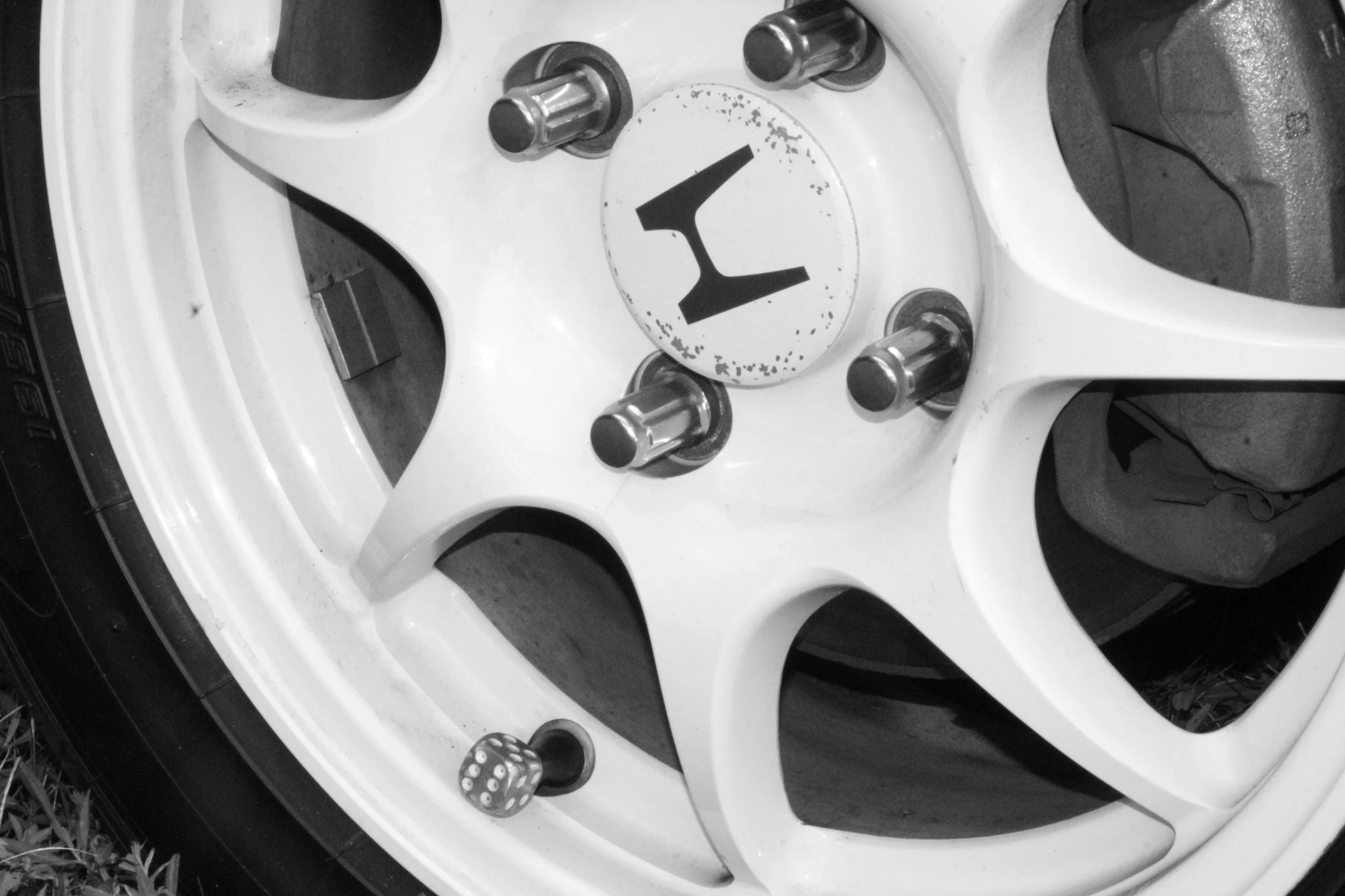 Close-up of a white car wheel with a hubcap that has the letter 'L' on it, surrounded by four lug nuts, with a small die showing a five on top and a black object at the top right.