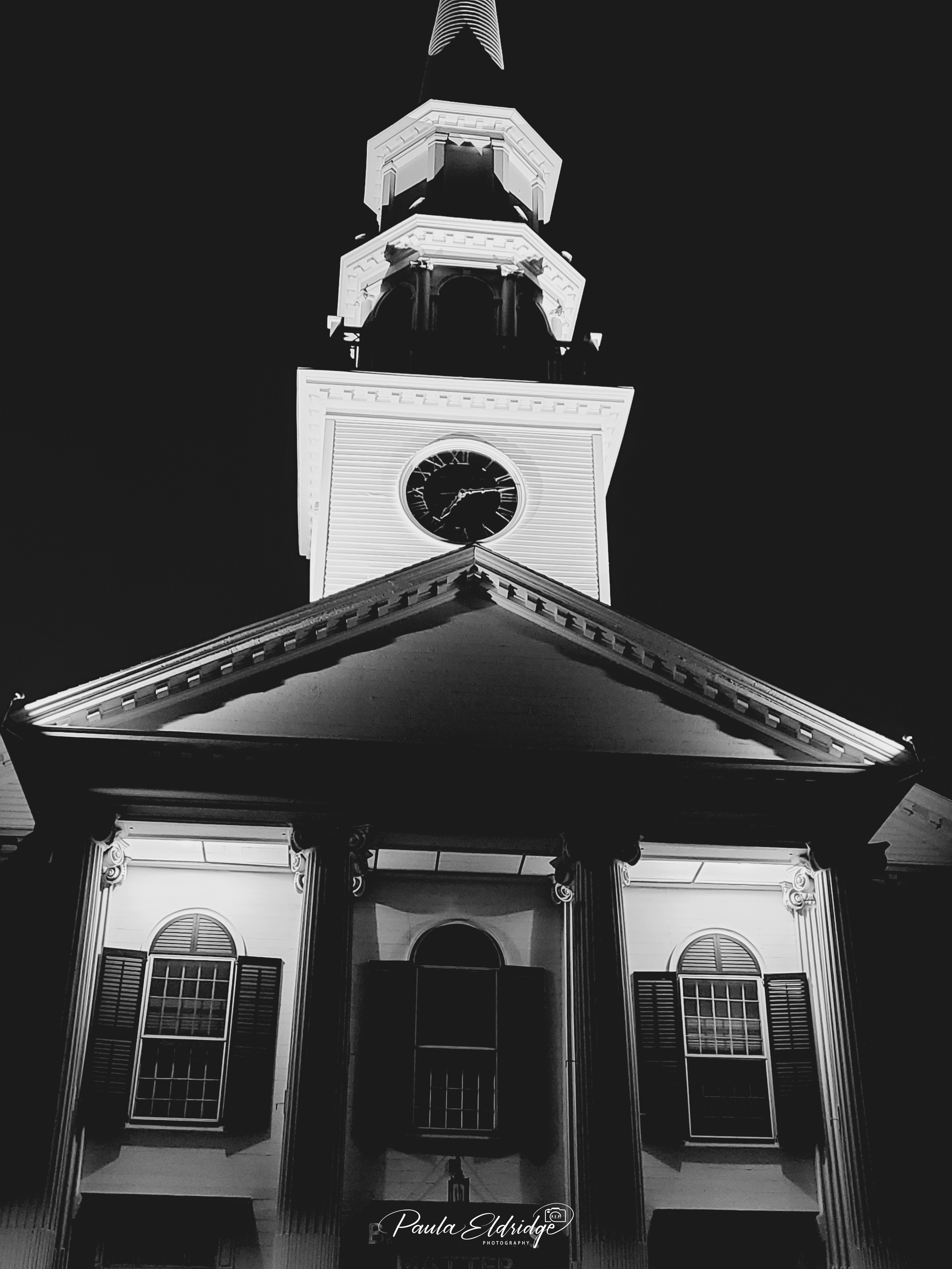 Nighttime view of a historic clock tower illuminated, showing the time, with a front of the building featuring columns, windows with shutters, and a triangular pediment.