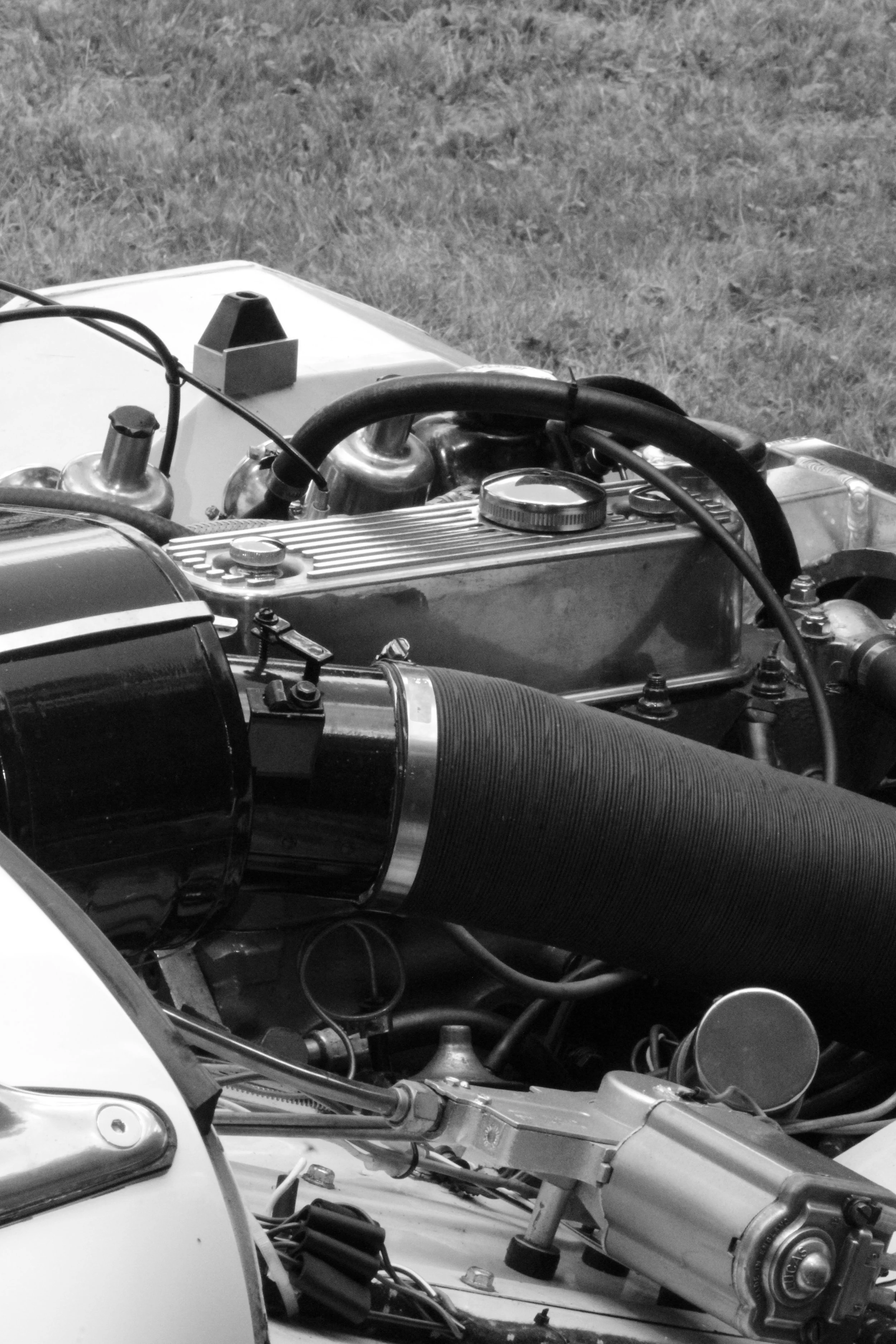 Close-up of a vintage car engine with various mechanical parts, hoses, and metal components visible, set against a grassy background.