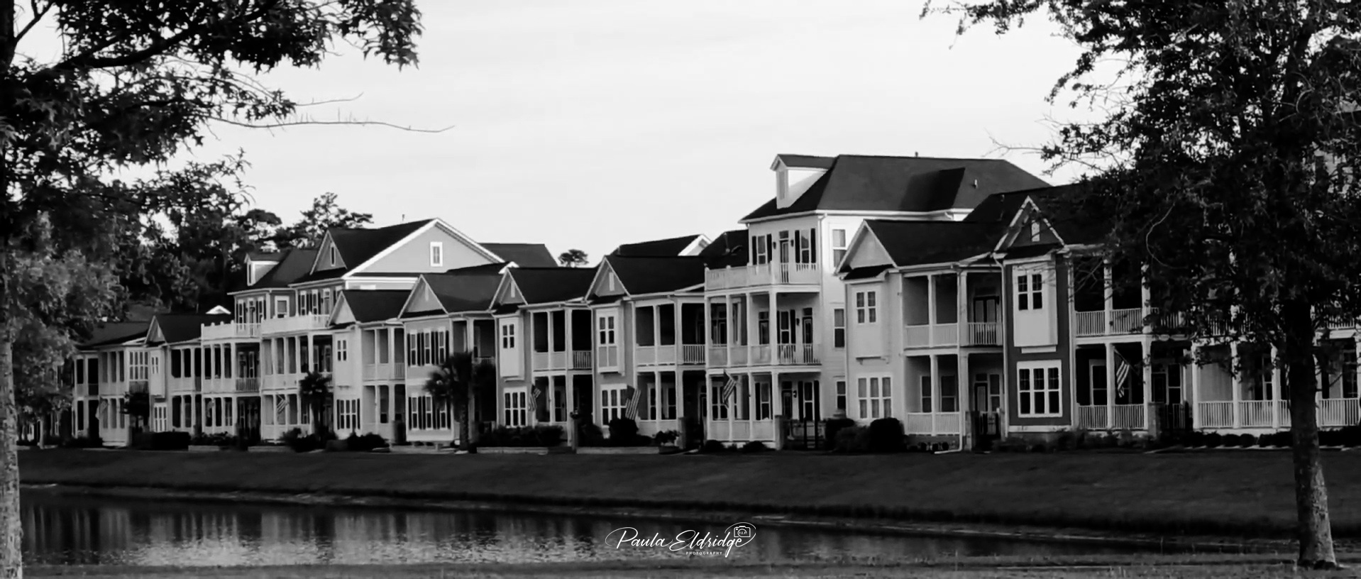 Black and white image of a row of modern multi-story houses with porches, situated along a body of water, with trees on either side.