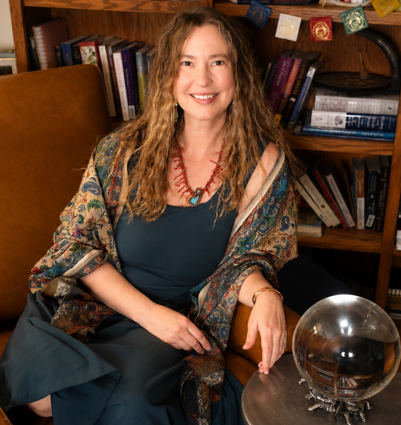 A woman with long curly blonde hair and a warm smile sitting in a cozy library, surrounded by books on wooden shelves, wearing a patterned shawl over a dark blue dress, with a glass orb on a stand on a wooden table beside her.