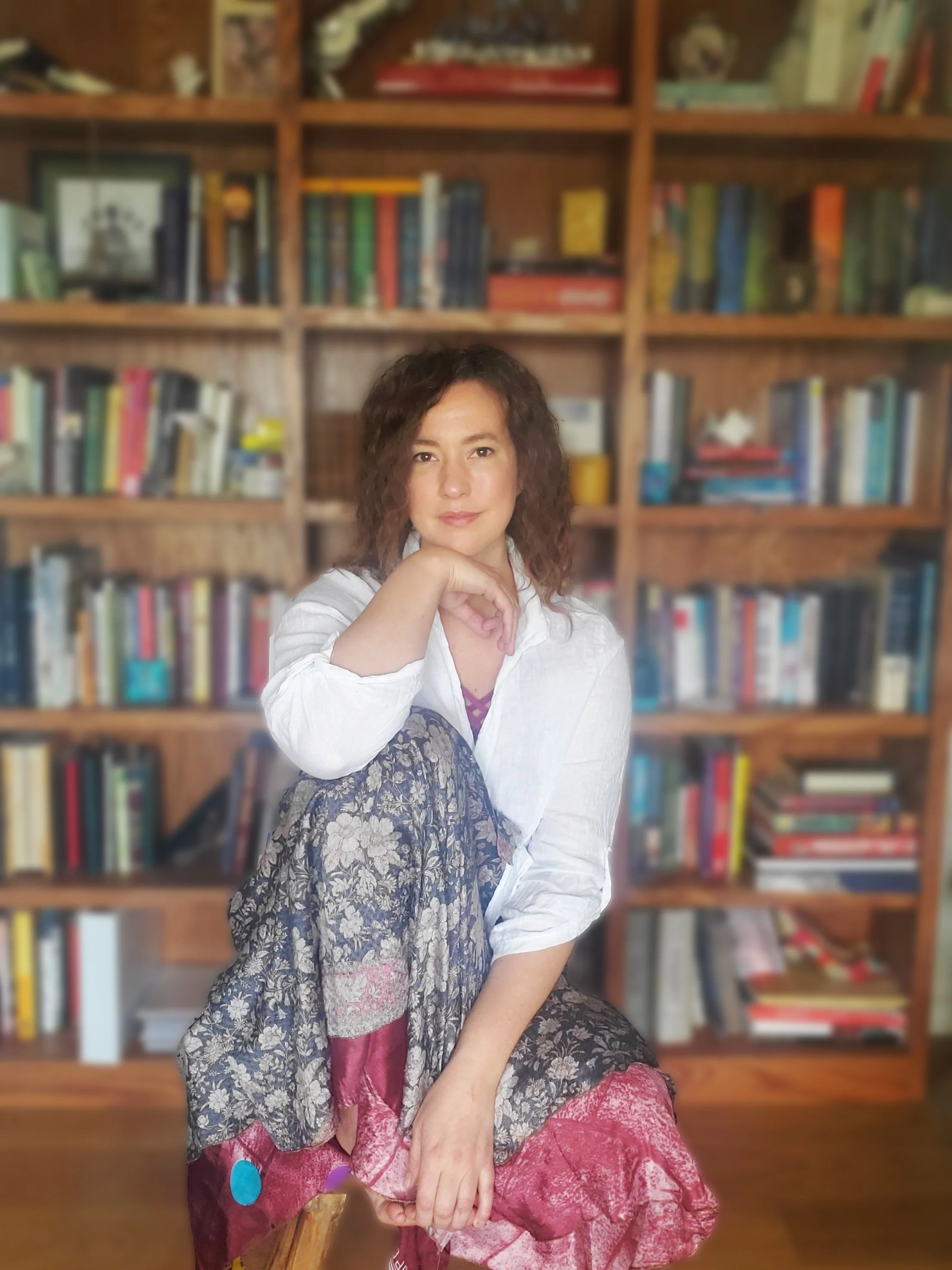 Woman with curly hair sitting in front of a wooden bookshelf filled with various books, wearing a white shirt and patterned skirt.